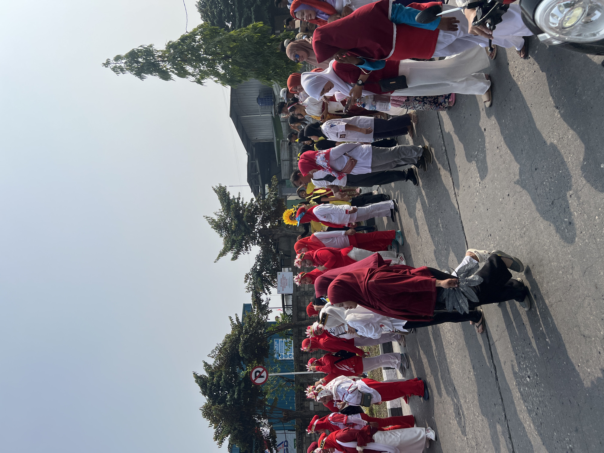 This image captures a daytime outdoor gathering of people in Klapanunggal, Indonesia, likely a parade or community event. A significant number of individuals are dressed in red and white attire, with many wearing headscarves, suggesting a cultural or celebratory occasion. Some participants are adorned with decorative headpieces. The group is standing on an asphalt road, with trees and buildings lining the background, indicating an urban or semi-urban environment. Shadows cast on the road suggest it is a sunny day. The presence of a "No Parking" sign points to a public street location. A motorcycle is partially visible in the foreground, hinting at the mode of transportation or presence of onlookers.