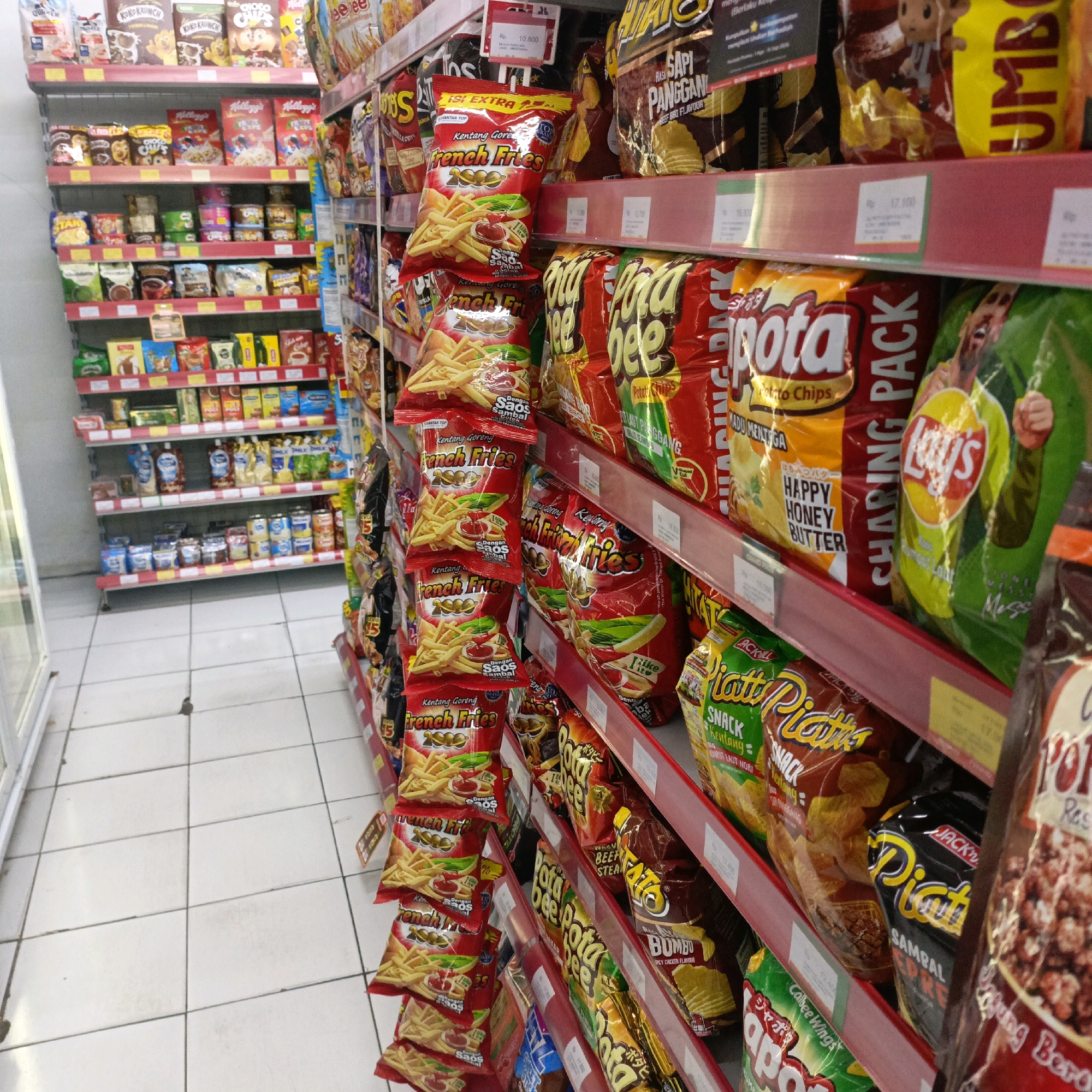 The image depicts a brightly lit interior of a minimarket, likely in Salatiga, Indonesia, as evidenced by the Indonesian language on product packaging and price tags. The scene is dominated by shelves stacked high with a variety of packaged snacks and food items, primarily potato chips and cereal.
On the right side of the image, several shelves are filled with colorful bags of chips. Prominently displayed are brands like "Pota Bee," "Lay's," and "French Fries" branded snacks, some with Indonesian text indicating flavors such as "Sapi Panggang" (Grilled Beef) and "Sambal" (Chili Sauce). Price tags are visible on some shelves, with currency indicated as "Rp" (Rupiah).
To the left, a separate aisle of shelves showcases more packaged goods. These include cereal boxes with cartoon characters, smaller snack bags, and bottles of sauces or condiments. The flooring is tiled with a light-colored grid pattern. The overall impression is one of a well-stocked retail environment focused on convenience foods. There are no people visible in the image, and the focus is solely on the product display.