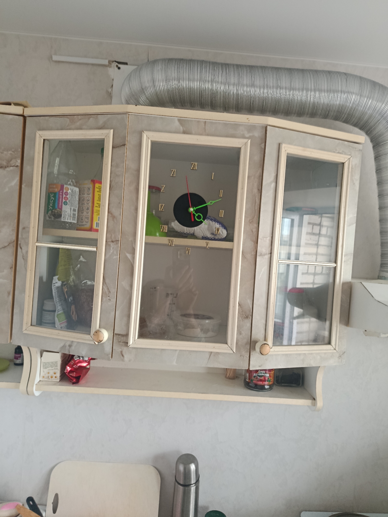 The image shows a kitchen scene, likely in Syzran, Russia, focusing on a wall-mounted cabinet and a shelf below it. The cabinet has glass doors revealing various items inside, including food products and a clock with Roman numerals. Above the cabinet, a large, flexible metal duct is visible, suggesting it's part of a ventilation system. The shelf below the cabinet holds a few containers, including a jar of sauce. The overall environment appears to be domestic, with a focus on storage and functional elements of a kitchen.