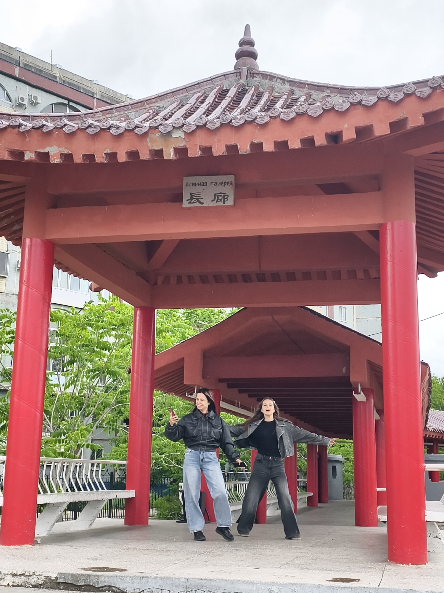 Two young women stand under a traditional Chinese-style pavilion with red columns and a tiered roof, located in Blagoveshchensk, Russia. The pavilion has a sign with Chinese characters above the entrance. The woman on the left is holding a red mobile phone and wearing a black leather jacket and light-wash wide-leg jeans. The woman on the right wears a gray jacket, a black top, and dark wide-leg jeans, with her arms outstretched. The background includes buildings and greenery, indicating an urban park setting.
