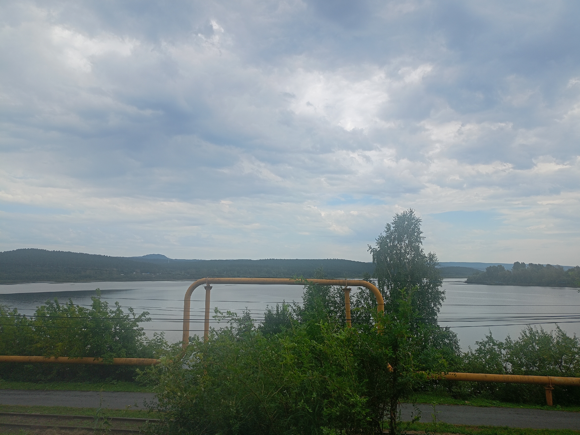 The image shows a wide, calm body of water, likely a river or lake, with forested hills in the background. The sky is overcast with scattered clouds, suggesting a temperate climate. In the foreground, there is a section of yellow industrial piping and lush green vegetation, including trees and bushes. Electrical wires are visible strung across the scene, running parallel to the piping. The ground in the immediate foreground appears to be a mix of gravel and asphalt, with some railroad tracks visible on the left. The overall impression is one of an industrial or semi-industrial area bordering a natural landscape. This scene is consistent with views of the Ay River within the city of Zlatoust, Russia, particularly in areas where industrial infrastructure meets the riverbank, possibly near the Zlatoust Metallurgical Plant. There are no people or animals present in the image.