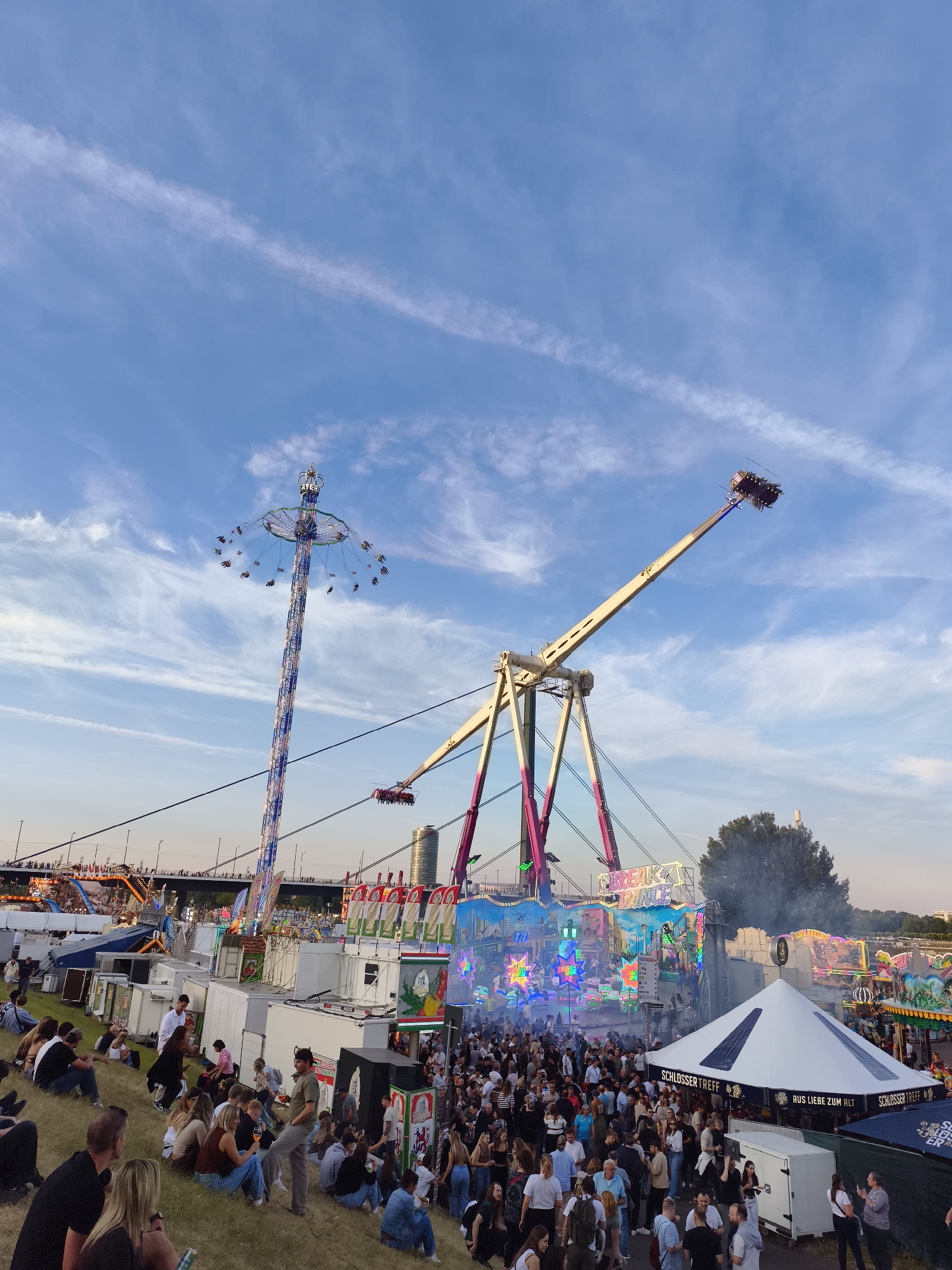 This is a wide-angle, eye-level photograph of a bustling fairground under a bright blue sky with scattered clouds. The scene captures a lively atmosphere with several amusement rides and numerous people milling about.
Dominating the upper half of the image are two prominent rides: a tall, suspended swing ride with people seated in gondolas is on the left, and a large, pendulum-like ride with seated passengers is on the right, angled across the frame. Both rides appear to be in motion. In the background, a bridge spans across a body of water, with city buildings visible in the distance, suggesting an urban setting.
The lower half of the image shows a large crowd of people gathered on a grassy slope, facing towards the fairground. They are dressed in casual attire. Several tents and stalls are visible, some decorated with colorful lights and signage. One tent in the foreground has the text "SCHLOSSER TREFF" and "AUS LIEBE ZUM ALT" visible. Smoke or steam is emanating from behind one of the attractions, adding to the dynamic feel of the scene.
The location is identified as the Rhein-Uferpromenade in Düsseldorf, Germany, which is a popular waterfront area often used for events and festivals. The time of day appears to be late afternoon or early evening, given the warm lighting and the angle of the sun.