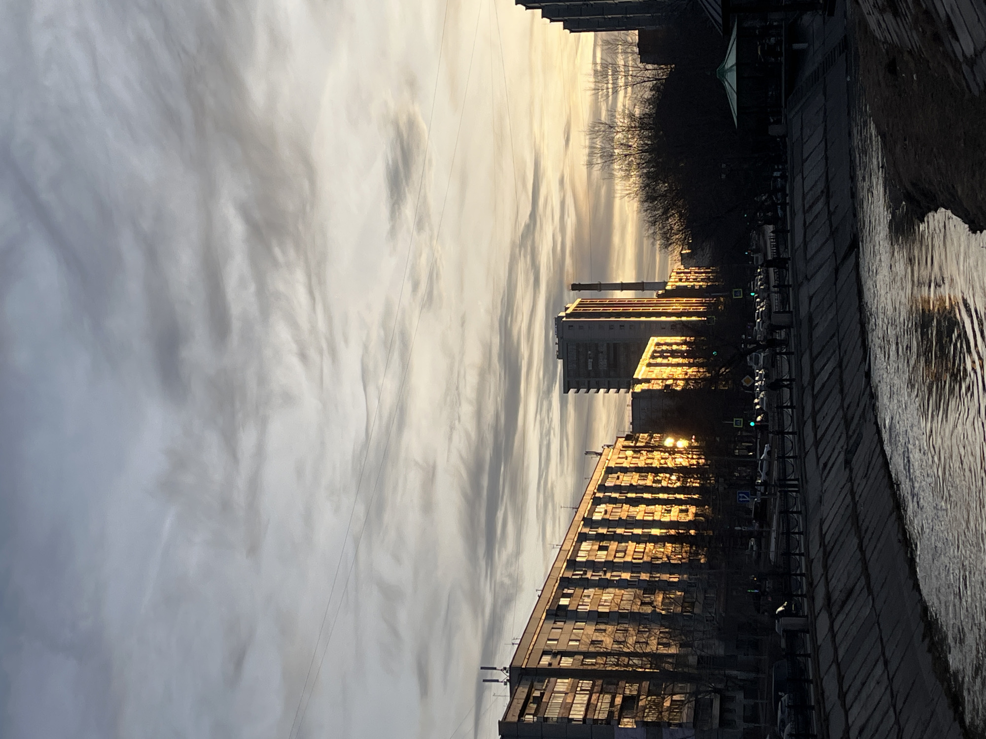 The image depicts a cityscape at dusk. The sky is overcast with striated clouds, reflecting a pale yellow light near the horizon. Multiple buildings, possibly apartment blocks, line a waterway, with the sun reflecting off their windows. There are several buildings and one apartment block has a dark, metallic sheen. A row of trees and shrubs runs along the edge of the walkway next to the waterway, with some appearing bare of leaves. Cars and streetlights are visible along the waterway. The photograph is rotated 90 degrees. It is most likely taken in Krasnoyarsk, Russia as it shows the city’s buildings and the Yenisey River.
