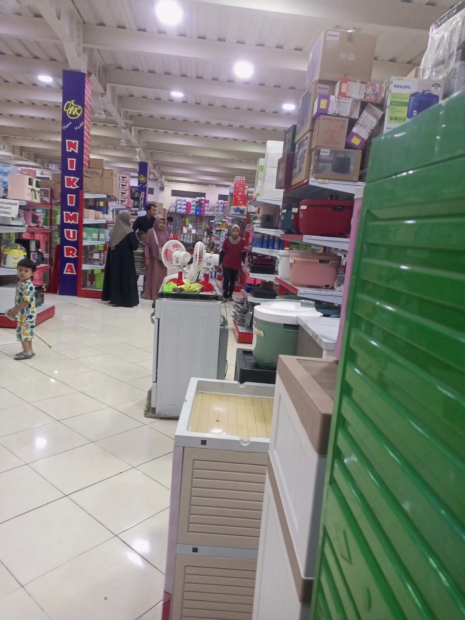 This image depicts the interior of a department store, possibly a "Niko Market" as suggested by signage. The store is brightly lit with fluorescent lights on the ceiling and features aisles stocked with various household items. In the foreground on the right, a large green cabinet with slatted doors is prominently displayed. To its left are stacked plastic storage drawers, one with a beige exterior and lighter beige drawer fronts, and another white drawer unit.
In the middle ground, several people are present. A young child wearing a patterned shirt and shorts stands on the left, holding a thin stick. Further back, three individuals are browsing. One person wearing a headscarf and dark clothing, another in a pink headscarf and pink dress, and a man in a dark shirt. A white oscillating fan is visible on top of a white appliance, possibly a washing machine, near the center of the image. Shelves in the background are filled with boxes, plastic containers, and other merchandise, including boxes labeled "PHILIPS". The floor is tiled with a light-colored pattern. The overall impression is a busy retail environment.