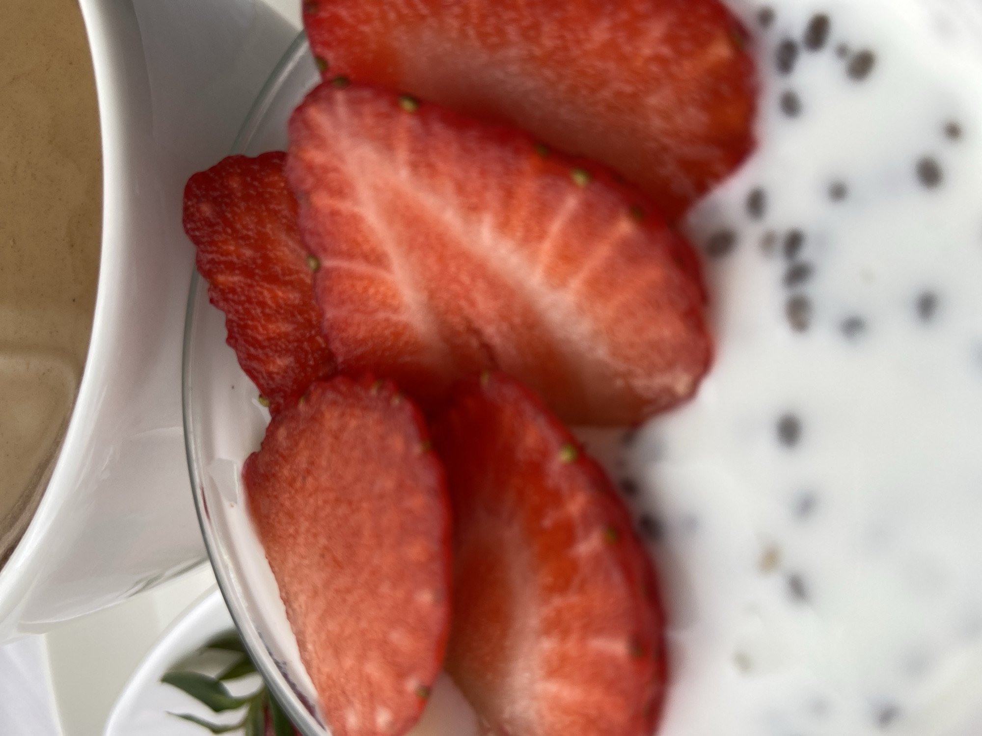 The image depicts a close-up view of a plate of food. Sliced strawberries are prominently displayed, partially overlapping the edge of the plate. Beside the plate appears to be a white pitcher, possibly containing a beverage or sauce. On the plate, a white substance with small, dark seeds is visible, suggestive of chia seed pudding or a similar dish. The background is blurred. The location in the city of Budva, Montenegro is not determinable from the image.