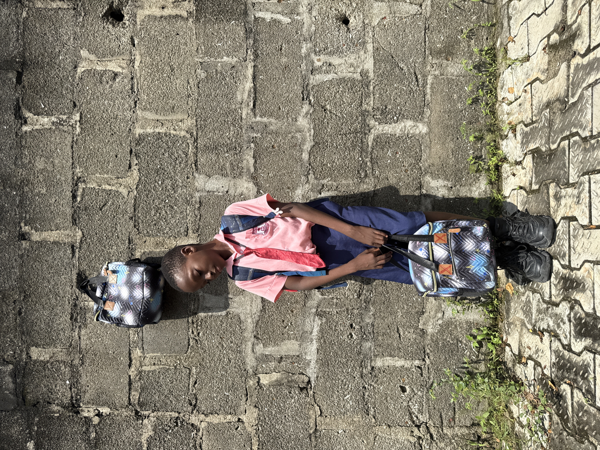 A young boy in a pink shirt, red tie, and blue shorts stands with his back to a rough concrete block wall in what appears to be Ado, Nigeria. He wears a backpack and holds a matching bag in his hands. Another backpack hangs from the wall to his left. The ground beneath him is paved with interlocking concrete blocks, and some sparse green vegetation grows at the base of the wall. The scene is illuminated by sunlight, casting shadows on the wall and ground.