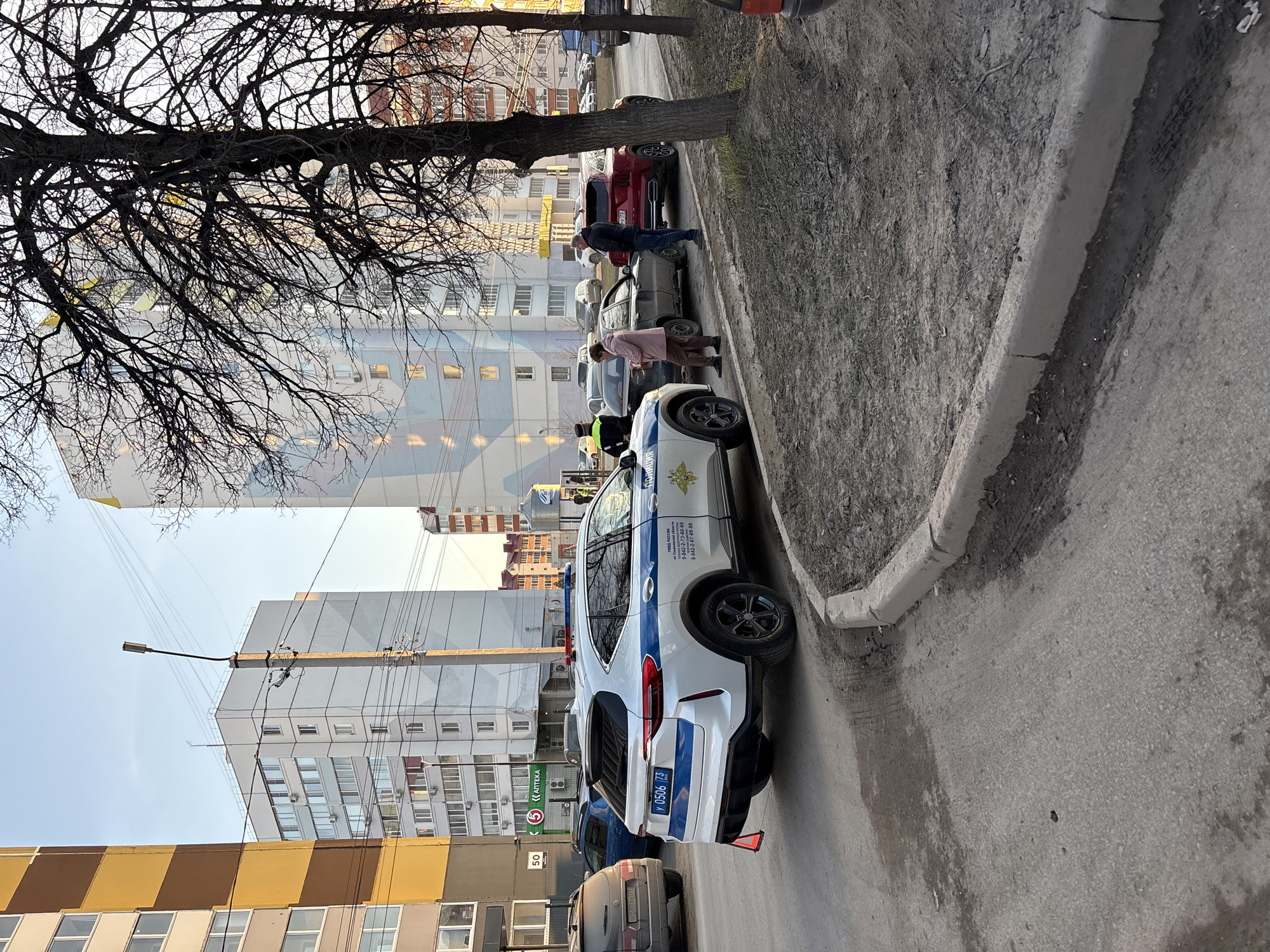The image captures an urban street scene in Ulyanovsk, Russia, likely during the daytime under a clear sky. The photo is taken from a slightly elevated perspective, looking down a street lined with buildings and parked cars.
In the foreground, a white police car with blue stripes is prominently parked on the side of a dirt strip next to a curb. The car bears the text "Полиция" (Police) and "УМВД РОССИИ по Ульяновской области" (Ministry of Internal Affairs of Russia for Ulyanovsk Oblast), along with contact phone numbers "8-842-2-73-60-60" and "8-842-2-67-88-88". A Russian coat of arms is also visible. The license plate reads "У 0506 73", confirming the Ulyanovsk region.
Several people are visible near the parked vehicles. A man in a dark jacket and a woman in a pink jacket are standing near a gray car, positioned between the police car and a red SUV further down the street. Other individuals are partially visible around the police car and further back, suggesting some activity or interaction, possibly related to a traffic stop or a casual gathering.
The street features a paved road next to a dirt shoulder and a curb. Overhanging bare tree branches dominate the upper left portion of the image, indicating a colder season. In the background, several multi-story buildings of varying architectural styles are present. One building has a distinctive checkered pattern in yellow and brown, while another displays a green "5 АПТЕКА" (Pharmacy) sign. A "50" speed limit sign is also visible on a pole. A streetlight and numerous power lines crisscross the sky above the buildings. The overall scene depicts a typical city environment with an official presence.