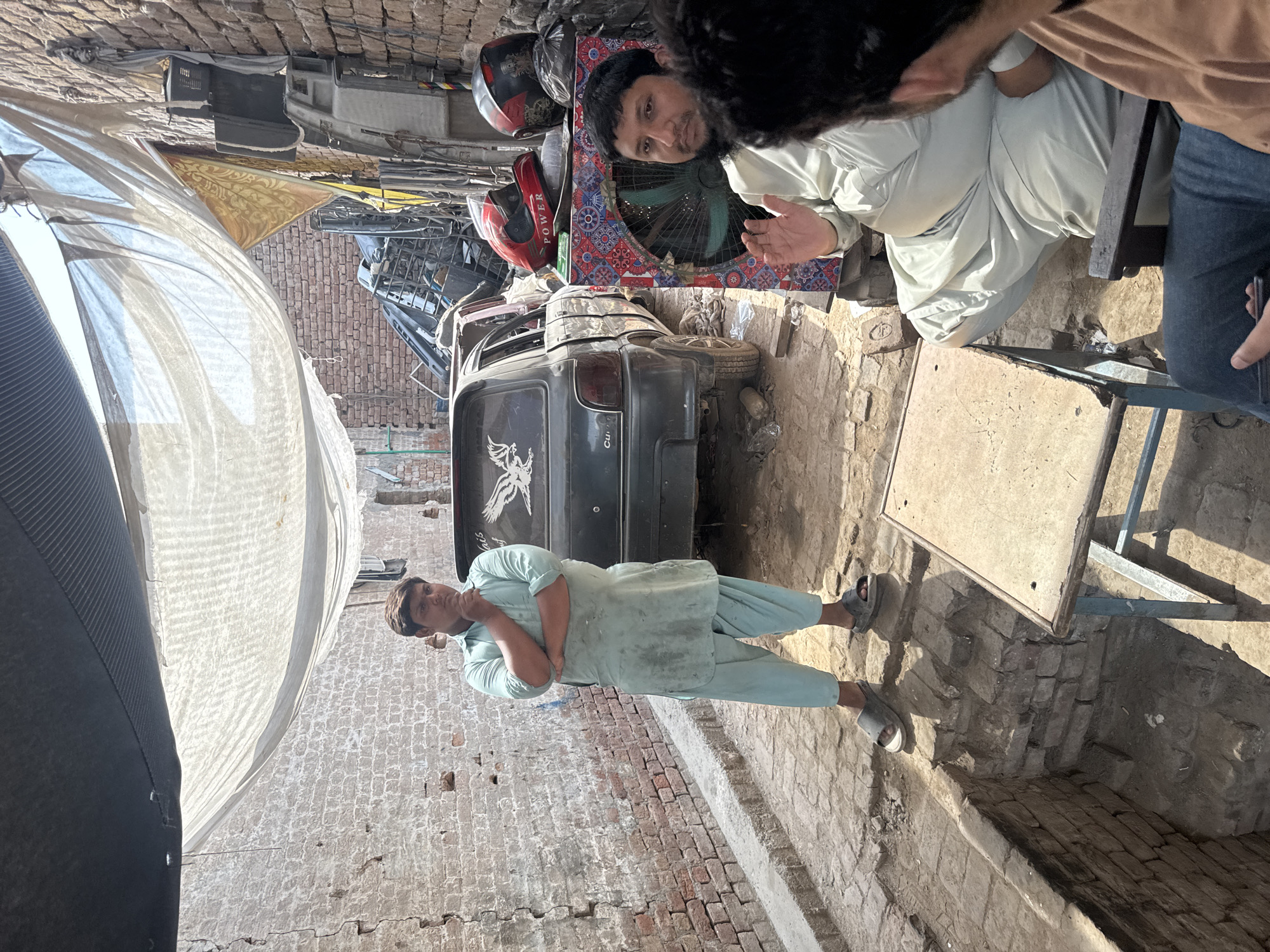 The image depicts a scene in Ghareeb Abad, Pakistan. Two men are present, one wearing a light green shalwar kameez with his arms crossed and the other is sitting next to a small table in the background and is obscured, wearing a light-colored shirt and jeans. A dark grey Suzuki car is parked in the background with a white decal on the back. Debris, including helmets and car parts, surrounds the vehicle. The environment appears to be an open-air mechanic shop or storage area with brick walls and varied ground surfaces. A makeshift awning provides partial shade.