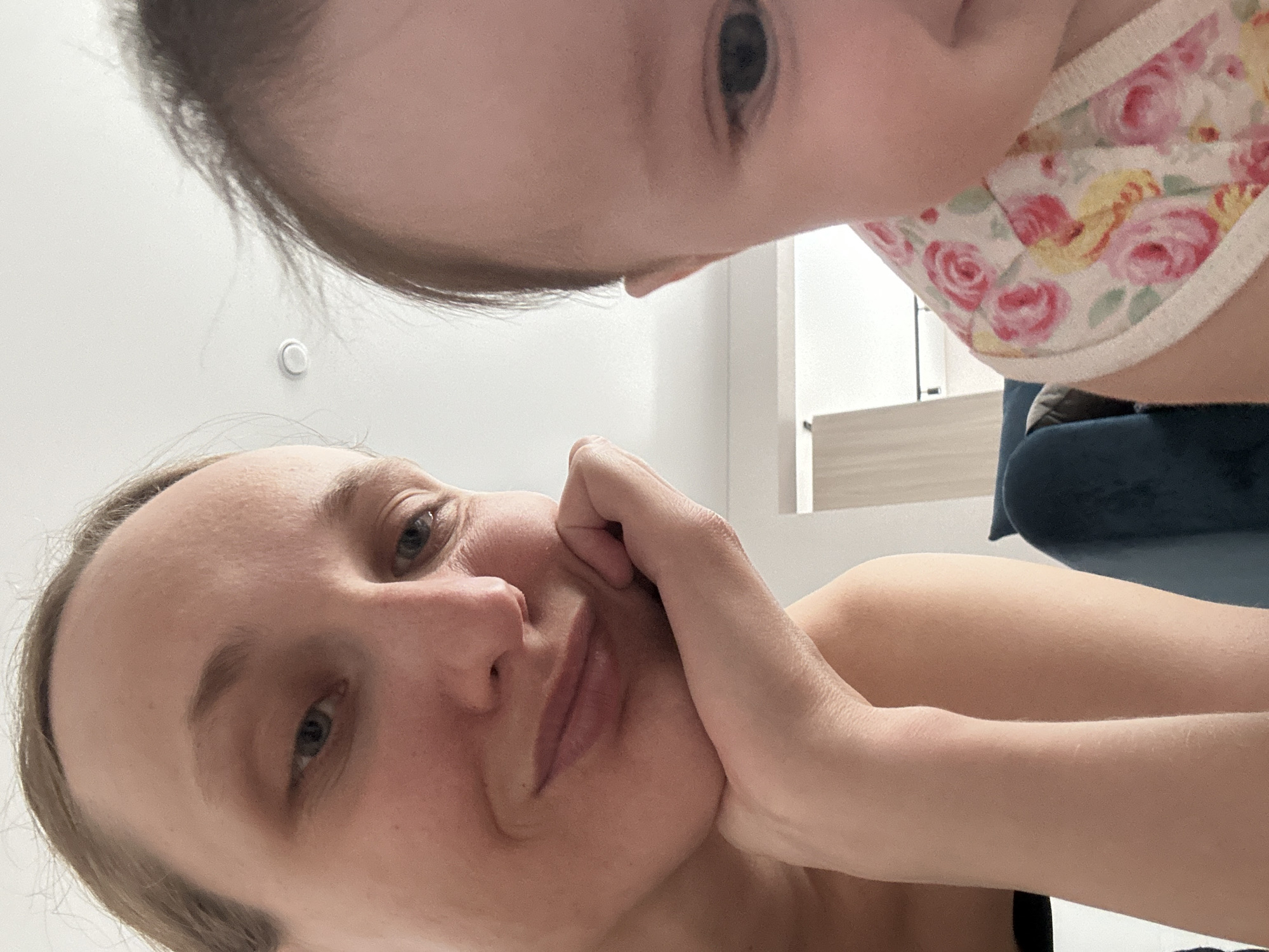 The image captures a close-up perspective of a woman and a young child, possibly in an indoor setting. The woman is looking at the camera with a hand resting on her face. The child is close to the camera with part of their face visible. A floral patterned bib is visible on the child. The background shows a neutral-colored wall and an edge of furniture. The image does not provide enough information to determine the location as Подшивалово, Russia.