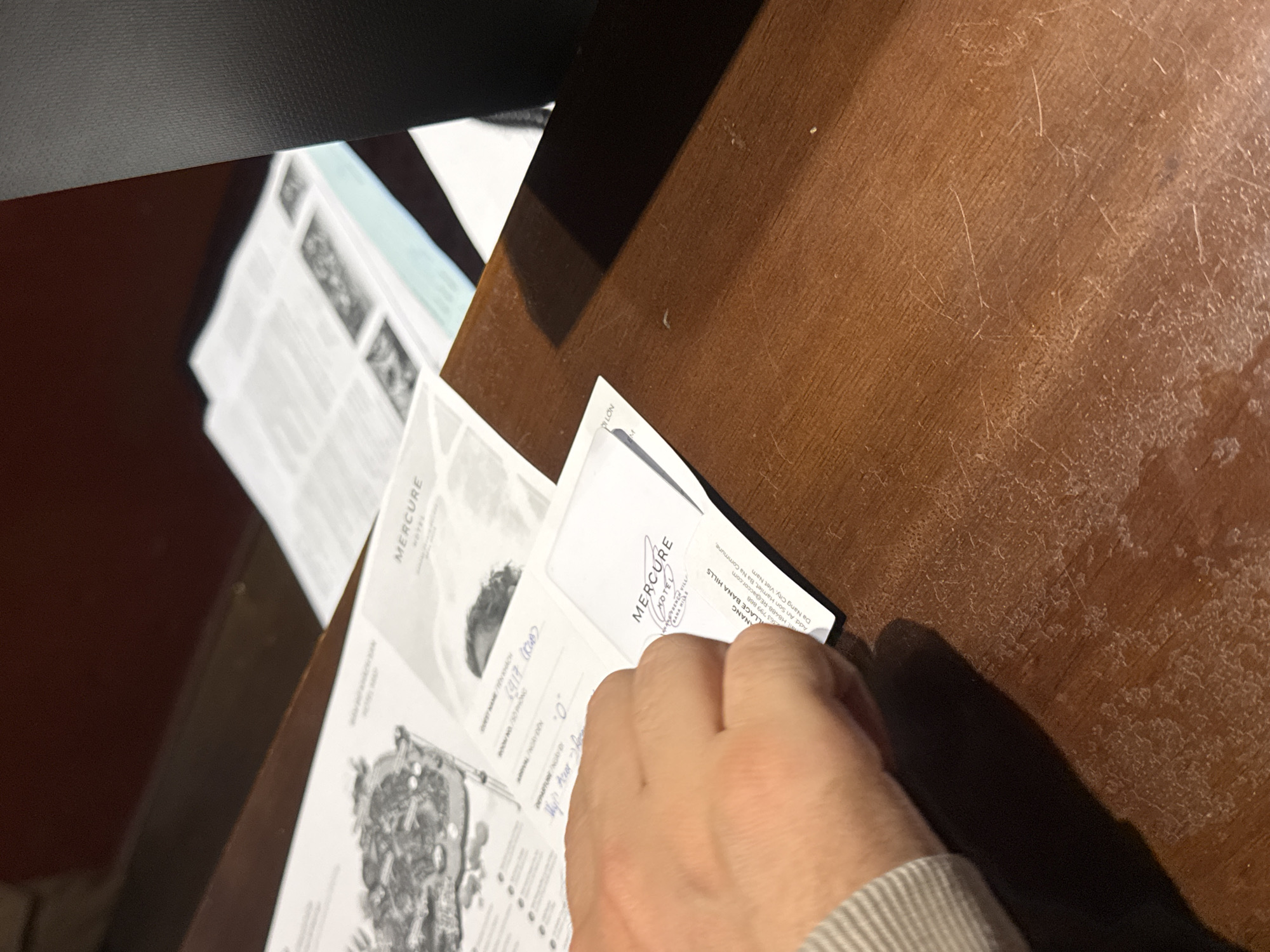 The image shows a close-up, angled view of a person's right hand holding or interacting with several white documents on a worn wooden table. The scene is indoors, likely at a hotel reception or desk. The wooden table is reddish-brown with visible scratches, scuff marks, and a layer of dust. A dark, textured surface is partially visible in the upper left corner.
The activity depicted is the handling of hotel check-in or information papers. Notable details confirm the location as a Mercure Hotel, specifically the "MERCURE HOTEL BANA HILLS" in Đà Nẵng, Vietnam.
Visible text on the documents includes:
* "MERCURE HOTEL" (repeated)
* "BANA HILLS" (repeated)
* "QUEST NAME/TEN KHÁCH"
* "6917 (100)"
* "ROOM NO/SO PHONG" and "0"
* Contact number: "363 799 868"
* Email address: "LH8488-RE@accor.com"
* Full address: "Add: An Son Hamlet, Ba Na Commune, Village Ba Na Hills, Da Nang City, Viet Nam"
* Partial words like "OLLON" and "RENEN VILL".
The image does not provide information about the time of day, weather, or the person's emotions.