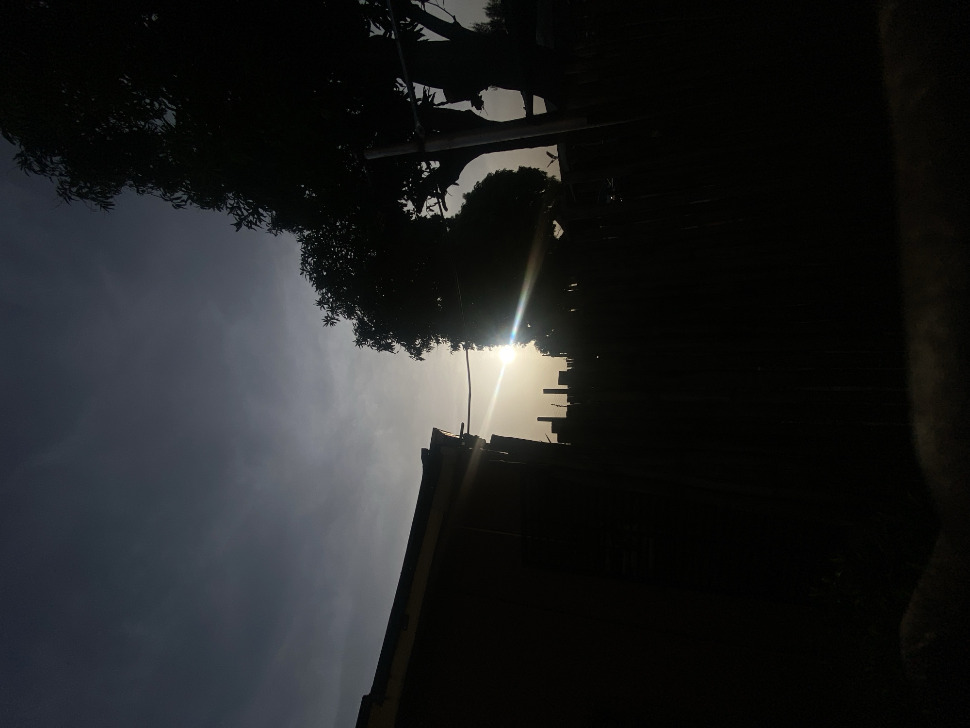 The image shows a low-angle, poorly-lit view of a partly cloudy sky above a building and trees. The building is silhouetted against a bright light source, presumably the sun, that is partly obscured by the building's roof and vegetation. In the upper left and right corners, there are dark areas which seem to be silhouetted tree canopies. An electrical wire cuts across the scene in the upper center. Specific details identifying the exact location in Barden Yana, Nigeria, are not discernible from the image.