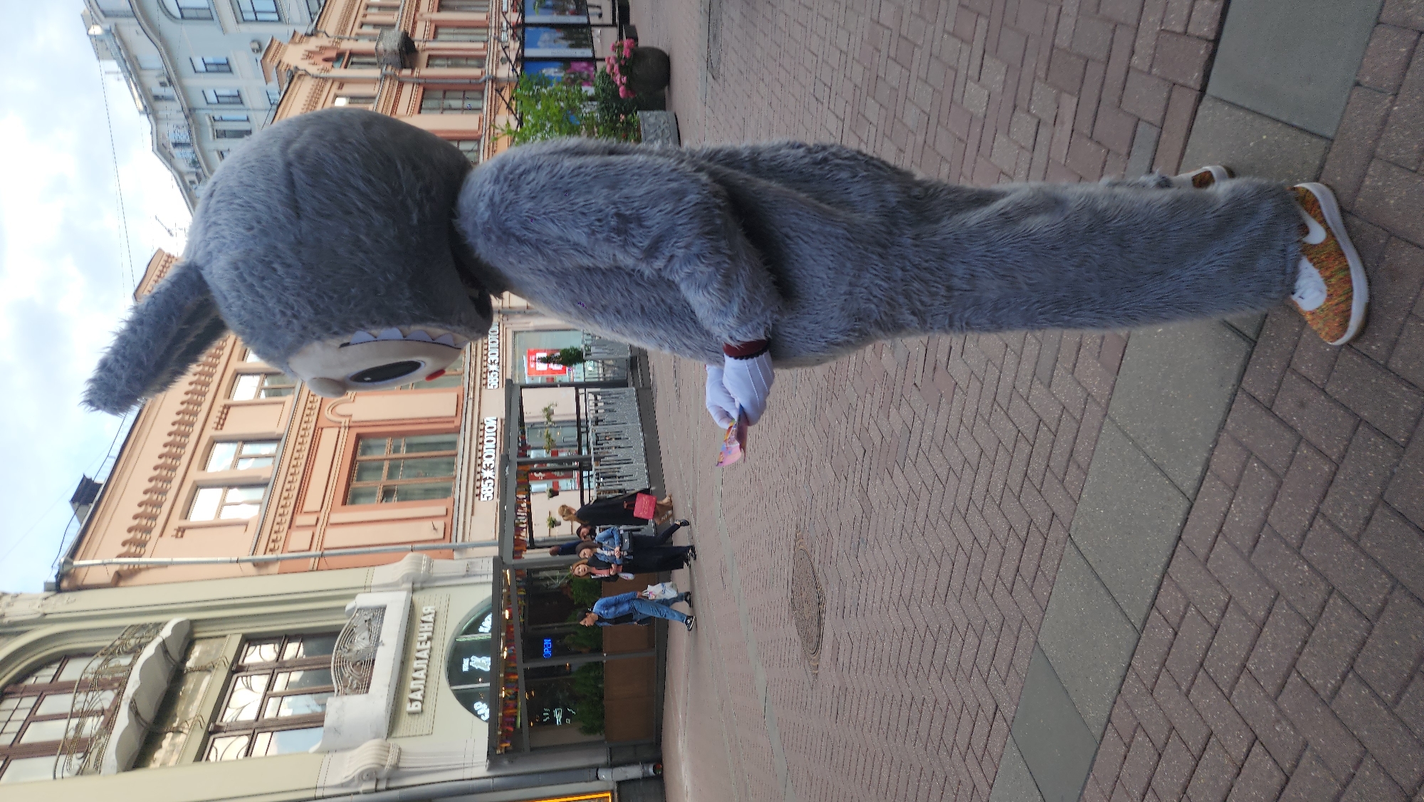 A full-body, side-view shot shows a person in a grey, fuzzy bunny costume standing on a brick-paved pedestrian street in Moscow, Russia. The costume has large ears, a round head with a cartoonish face, and large, fluffy body suit. The bunny's left hand, wearing a white glove with a red cuff, holds a folded piece of pink and yellow paper, possibly a flyer or coupon. The bunny is wearing colorful, speckled sneakers.
In the background, pedestrians walk past shops and cafes. To the left, a building with an ornate facade and balconies is visible, with a sign that reads "БАЛАЛАЕЧНАЯ" (Balalaechnaya). Further down the street, signs for "585 ЗОЛОТОЙ" (585 Zolotoy), a jewelry store chain, are prominent on other buildings. The sky is overcast, and the general atmosphere suggests an urban, commercial street. The pavement is laid with a herringbone pattern of red-brown bricks, with a grey strip of rectangular pavers running through the middle.