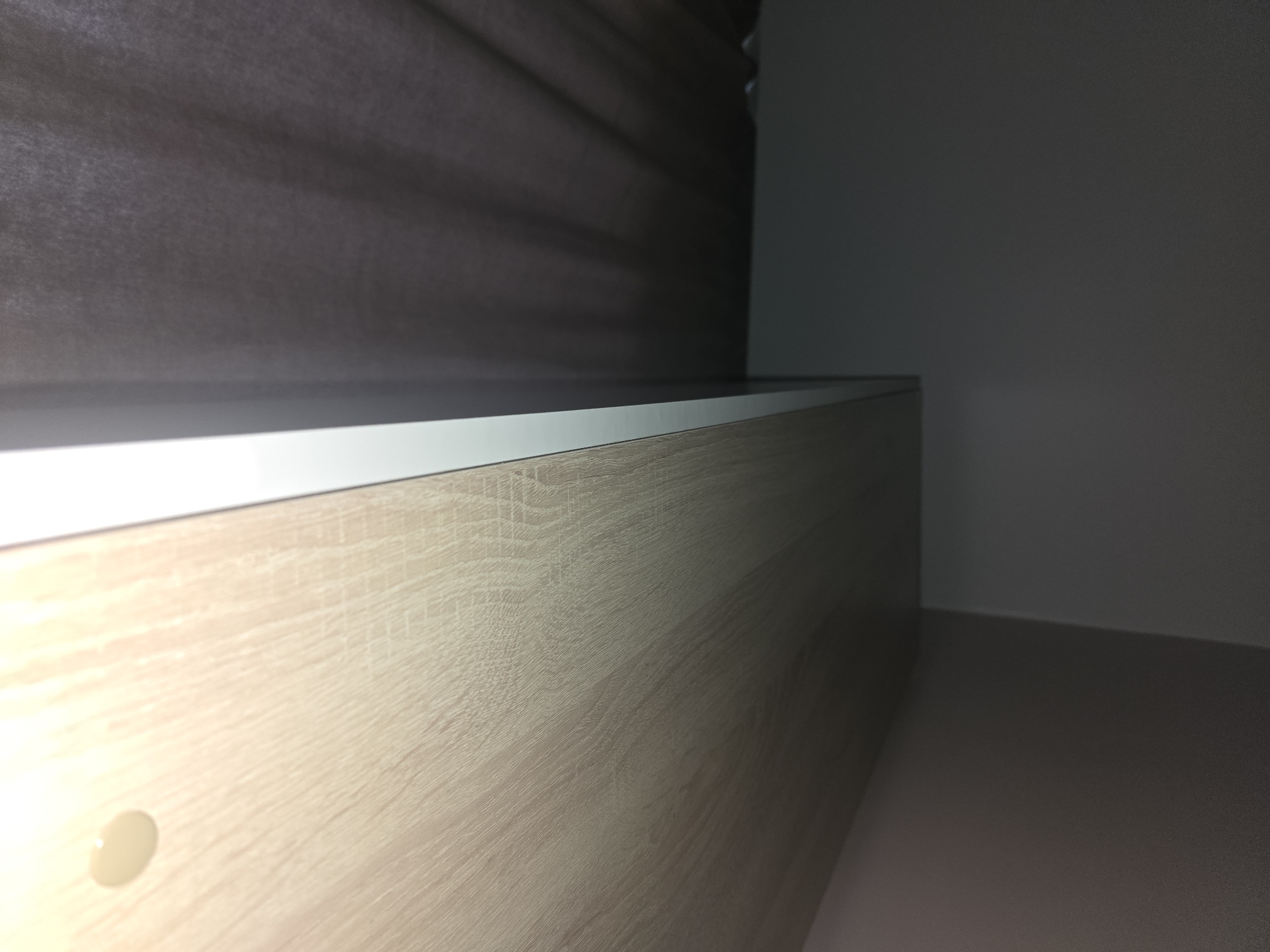 The image shows a close-up, eye-level, wide shot of a light-colored wooden surface with a wood grain texture. A white edge runs along the top of this surface. To the left, a dark gray, textured material, possibly fabric or a blind, is visible, with soft folds and shadows cast upon it. The background is dimly lit and transitions from a dark gray on the left to a lighter gray on the right, suggesting an interior space. There are no people or overt actions depicted. The location in Ulan-Ude, Russia cannot be determined from the image content.
