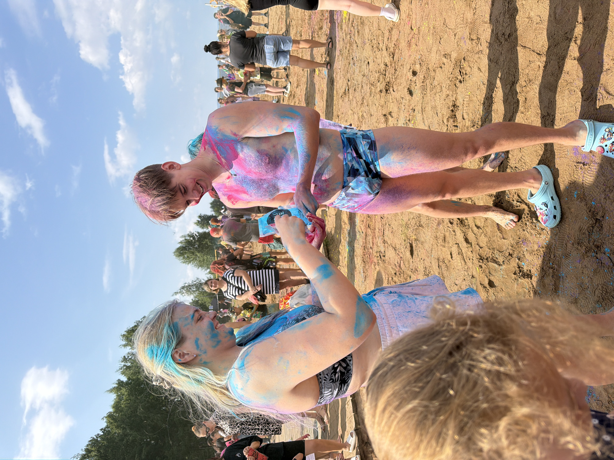 The image captures a lively outdoor event, likely a festival or celebration, set on a sandy, outdoor area. The ground is covered in sand and traces of colored powder, suggesting a Holi-like festival. In the foreground, a young woman with blonde hair, partially dyed blue, is interacting with a young man. Both are covered in vibrant pink and blue powder. The woman wears a black and white patterned bikini top and white shorts, while the man is in patterned swim trunks and light blue Crocs. Behind them, a crowd of people are visible, some also with colored powder on their faces and clothing, indicating a collective participation in the event. The background features a line of trees and a bright, partly cloudy blue sky. The overall atmosphere appears festive and joyful. The location is identified as the beach in Chaikovsky, Russia.