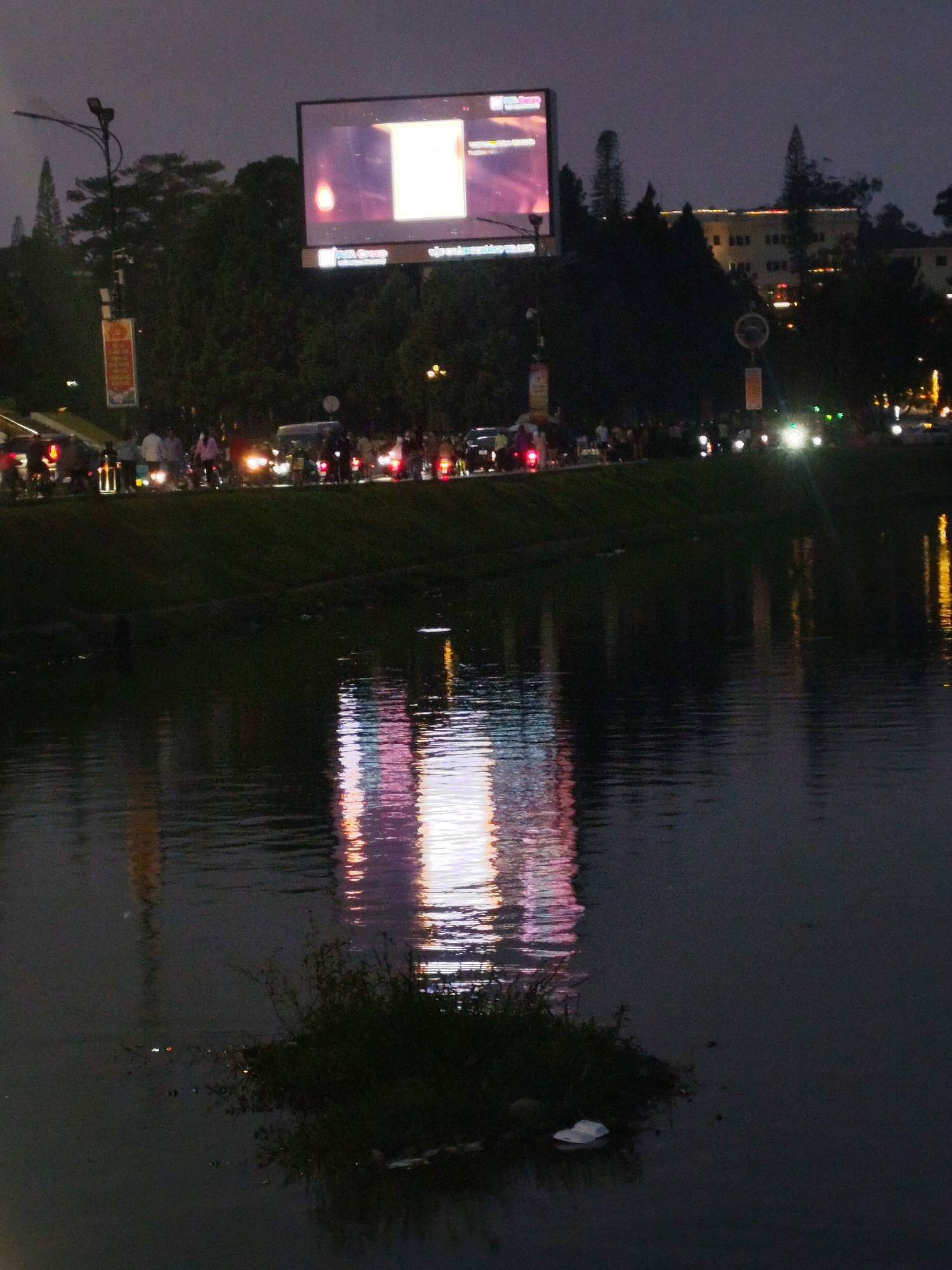 This is a nighttime photograph of a canal or river in Da Lat, Vietnam. A large LED screen or billboard is visible in the background, its bright light reflecting in the water. Traffic, including motorcycles and possibly cars, is visible along the road bordering the canal. The scene is dimly lit, with the primary light sources being the screen, vehicle headlights, and streetlights. There are trees and other vegetation in the background, adding to the urban landscape. An area of green vegetation protrudes from the water in the foreground.