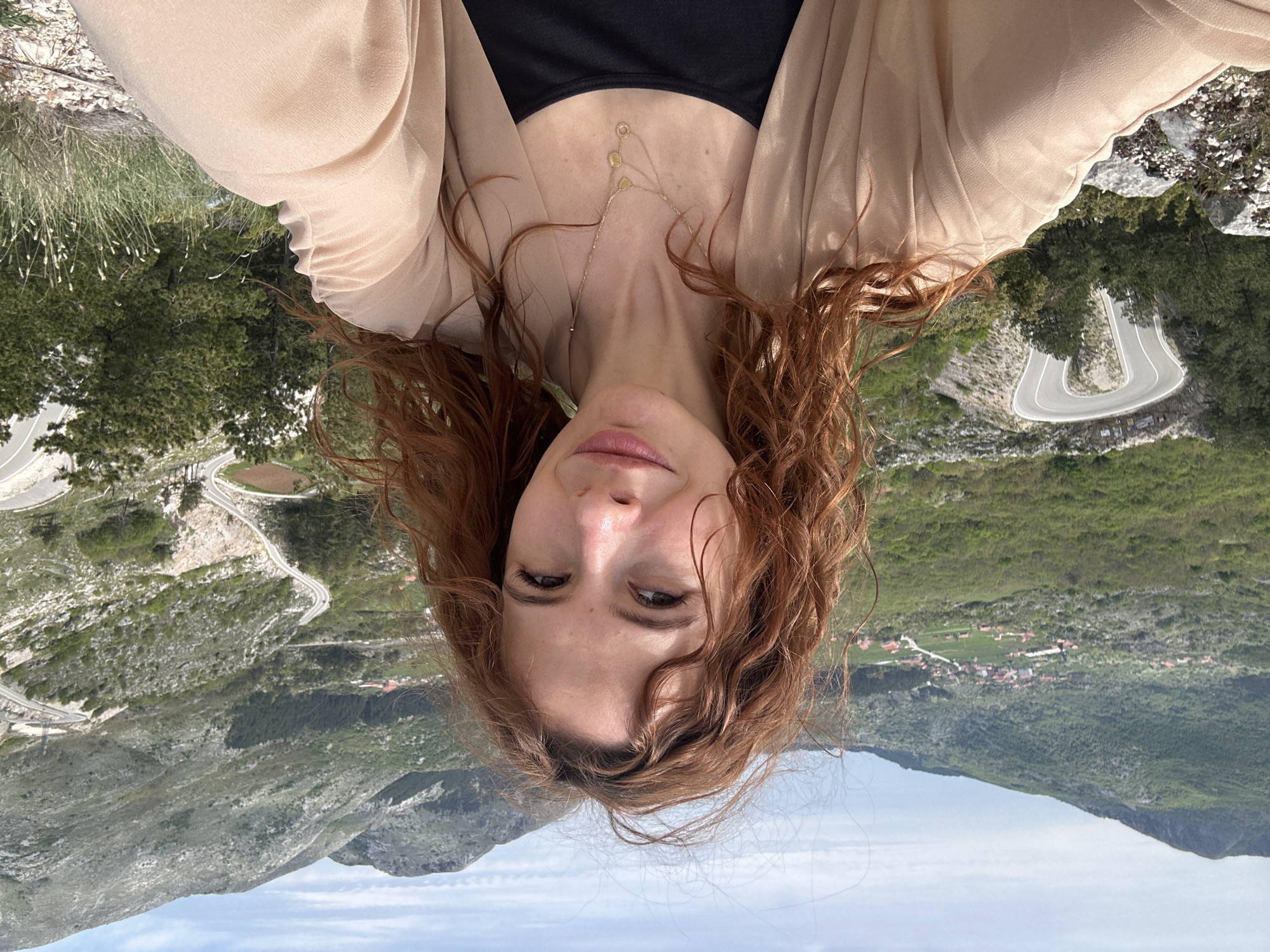 A woman with curly red hair, a black top, and a beige outer garment is pictured in a vertical composition against a mountainous backdrop. A gold necklace adorns her neck. The landscape shows green vegetation. In the city of Bukovica, Montenegro, a winding road is visible in the background.