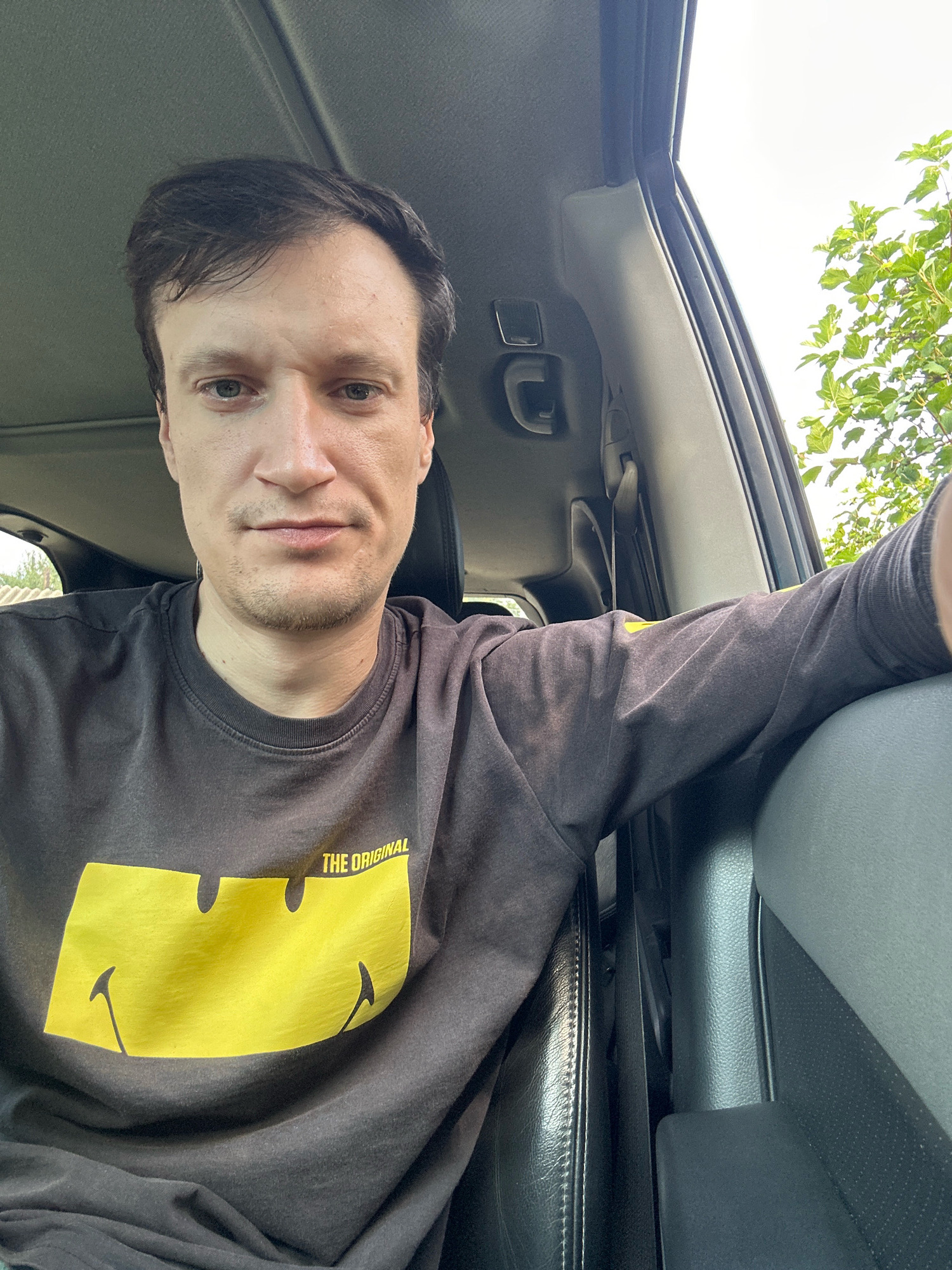 A man is captured in a selfie taken inside a car, with a large yellow smiley face and the words "THE ORIGINAL" printed on his dark gray sweatshirt. He has brown hair and is looking directly at the camera. The car interior is visible, including a gray seat and a portion of the car's roof. Outside the car window, green leaves from a tree are seen against a bright sky. The scene is likely located in Shatava, Ukraine, a town situated in the Chortkiv Raion of the Ternopil Oblast.