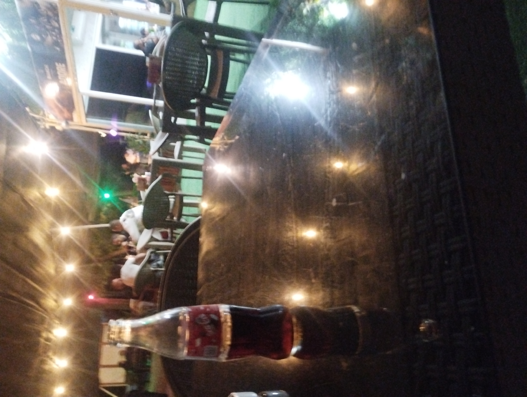 The image captures a dimly lit outdoor seating area, likely at a restaurant or cafe, with strings of lights illuminating the scene. In the foreground, a glass bottle of Coca-Cola rests on a dark, reflective table. The bottle is partially filled with the dark liquid, and its label is visible.
In the background, several people are seated at tables, engaged in conversation or eating. The setting appears to be a communal outdoor space, with thatched roofing or a similar covering overhead, supported by posts. A building with large windows and signage can be seen further in the background, with the word "ADORE" partially visible on a sign. The ground is covered with artificial grass. The overall impression is that of a relaxed evening gathering. The specific location within Karnobat, Bulgaria, is not discernible from the visual cues alone.