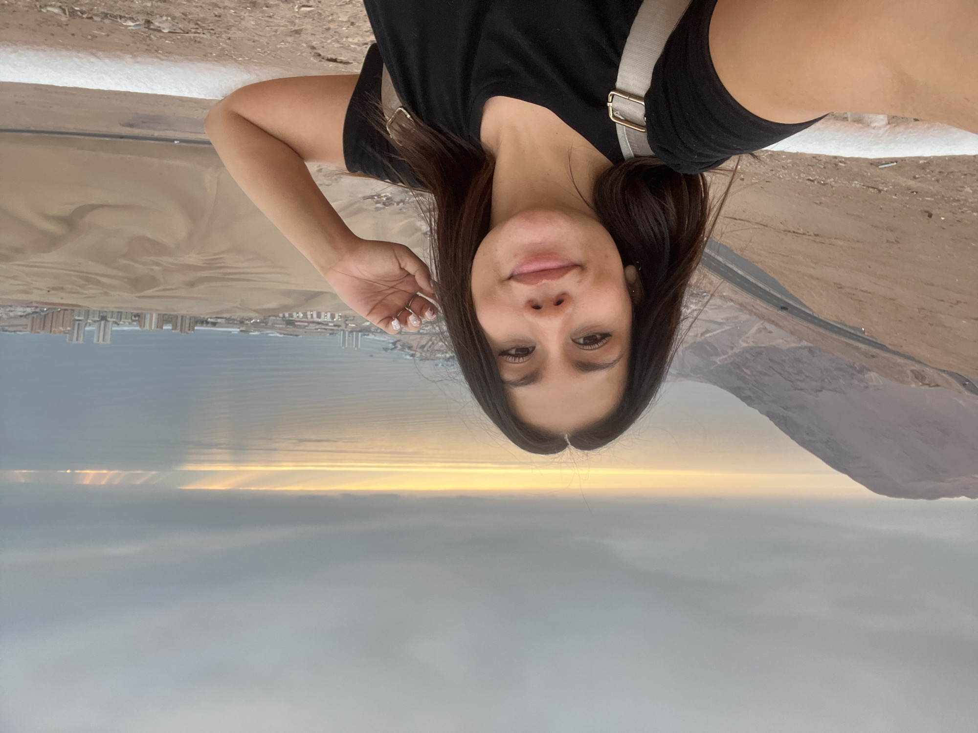The image shows a woman in a black t-shirt and shorts, with a light-colored strap across her chest, standing on a white ledge. She is fair-skinned with dark hair, and she is smiling. Her left hand is raised towards her hair.
Behind her, there is a coastal landscape with buildings, sand, and a road. The horizon line is visible with light shining through the clouds. On the right side of the image, there is a mountain range.
The location appears to be a coastal area, possibly near Iquique, Chile, given the desert-like terrain, ocean views, and city visible in the distance.