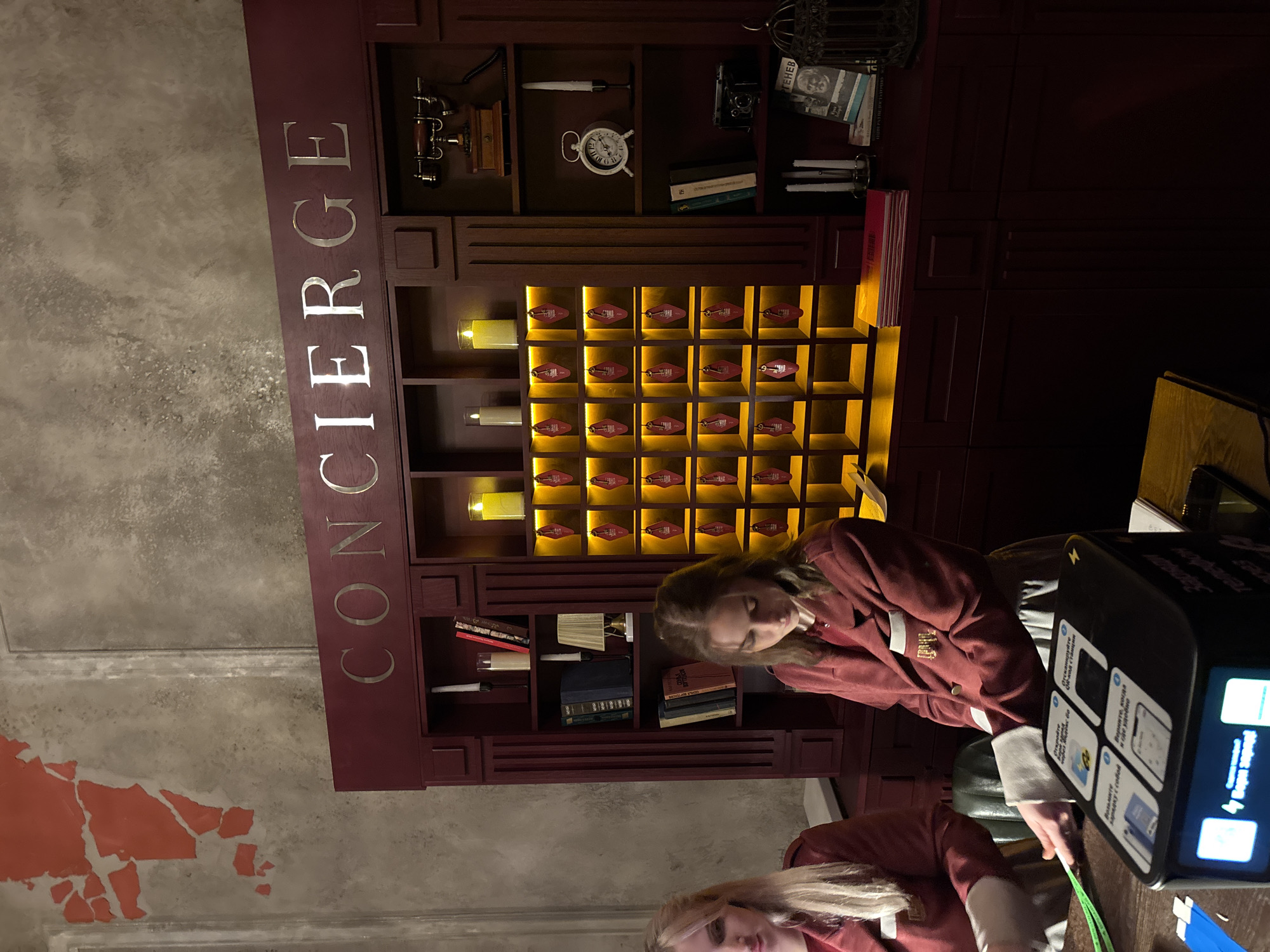 This vertical image depicts an indoor scene at a concierge desk, likely in Krasnoyarsk, Russia. The main subject is a large, maroon-colored wall unit with the word "CONCIERGE" prominently displayed vertically in metallic letters on its left side.
The unit's upper shelves are adorned with various vintage items, including an old telephone, a white antique clock, several books (one with "ГЕНЕВ" visible on its spine and another with "ТЕКНЕВ"), candles, and a small decorative cage. Below these shelves, a grid of illuminated compartments holds numerous key fobs, characteristic of traditional hotel key storage.
A young woman, dressed in a maroon jacket with light cuffs and gold embellishments (suggesting a uniform), stands at a dark desk in front of the unit, looking down. She appears to be interacting with a digital kiosk on the desk. The kiosk displays Russian text, including "Возьмите зарядку с собой" (Take charger with you), "Откройте... и где удобно" (Open... and where convenient), "Верните, когда" (Return when), and a partial "телеф" (phone). A clear reflection of the woman and the room is visible on the shiny surface of the desk or floor in the foreground.
The wall to the left features a textured, grey finish with sections of peeling red paint. The lighting is warm, with bright yellow light emanating from the key compartments, indicating an indoor setting, possibly during the evening. The overall ambiance combines classic and modern elements.