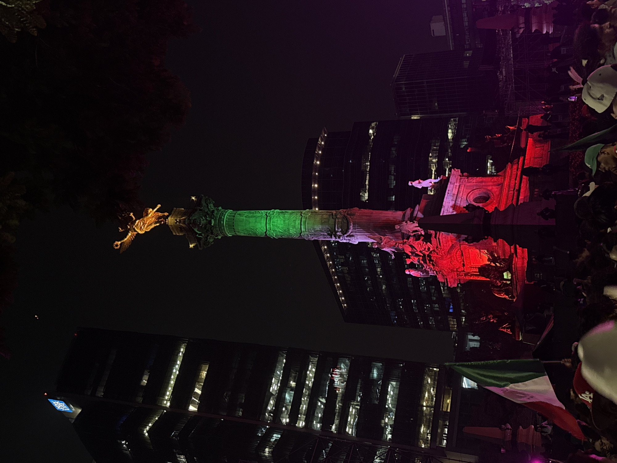 The image captures a night scene in Mexico City, likely near the Monumento a la Independencia (Angel of Independence). A tall, ornate monument is illuminated with green and red lights, resembling the colors of the Mexican flag. A golden statue of an angel stands atop the monument, holding a laurel wreath. Surrounding the monument are contemporary buildings, including a dark skyscraper with illuminated windows on the left. In the foreground, a crowd of people is visible, some holding a Mexican flag. The atmosphere appears festive, with colored lights and a gathering of people suggesting a celebration or event.