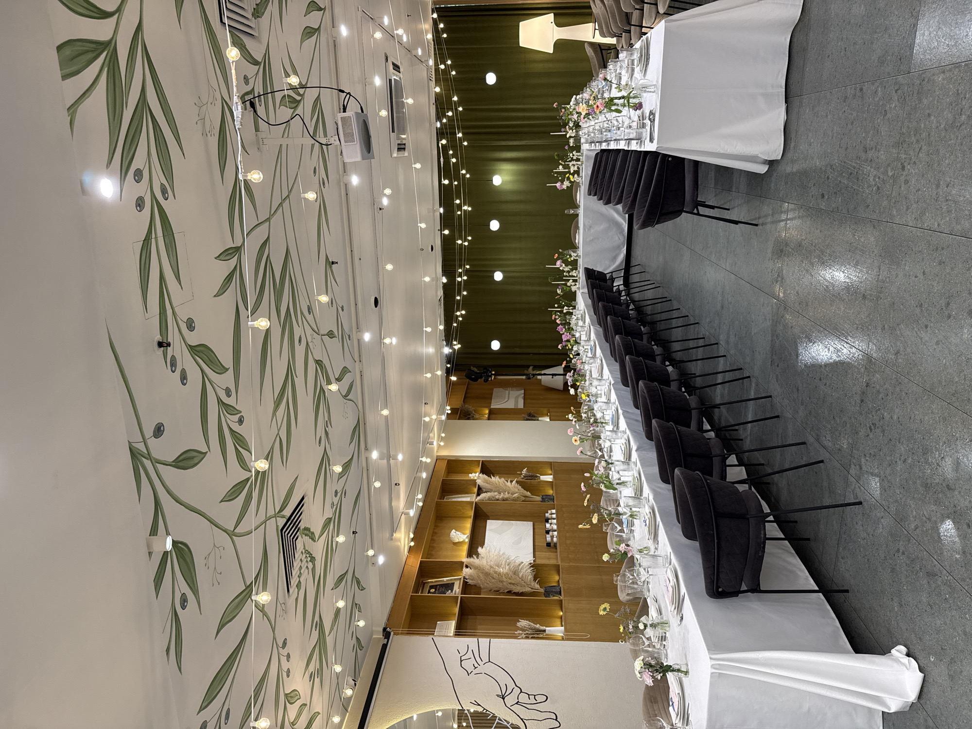 The image depicts an interior setting resembling a wedding reception or formal event venue, possibly located in Saint Petersburg, Russia (given the architectural style and prevalent decor).
The scene focuses on a long, rectangular table set for a banquet, draped with a white tablecloth and decorated with floral arrangements and glassware. Black chairs are arranged along both sides of the table. The floor is a gray, smooth material, perhaps tile or polished concrete.
The left side of the frame showcases a wall adorned with a large botanical mural featuring green leaves and berries on a white background. String lights are hung along the wall and ceiling, adding to the festive ambiance. Towards the end of the wall, a projection screen and projector are seen.
Adjacent to the end of the banquet table is a wooden display case filled with decorative objects and art pieces, including framed artwork and sculptures. A black and white drawing of a hand is painted on the wall beside the case.
A green curtain is visible in the background, suggesting a stage or performance area. Overall, the scene suggests a well-decorated space prepared for a special event.
