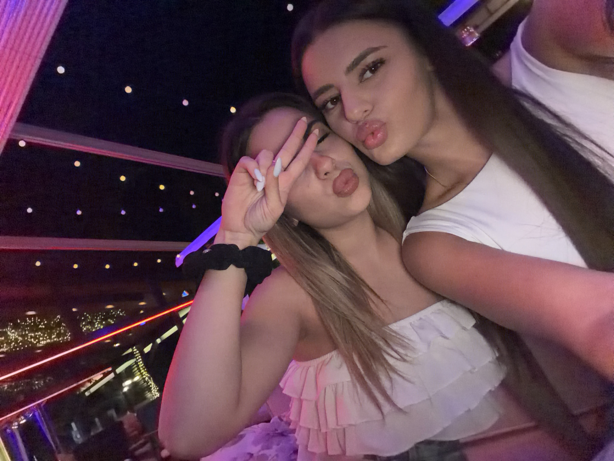 This is a close-up, selfie-style photograph of two young women, likely in their late teens or early twenties, posing for a picture. They are both wearing white tops and have their lips puckered, as if for a kiss. The woman on the left has her hand up, making a peace sign over her eye. The background appears to be dark, with some blurred lights that suggest an urban or entertainment setting, possibly at night. There are some red and pink light streaks visible, and a few scattered white and yellow lights in the background that could be from a cityscape or decorative lighting. The overall lighting is dim and atmospheric, with colored lights casting a glow on the subjects. The image does not contain enough information to identify the location as Haskovo, Bulgaria.
