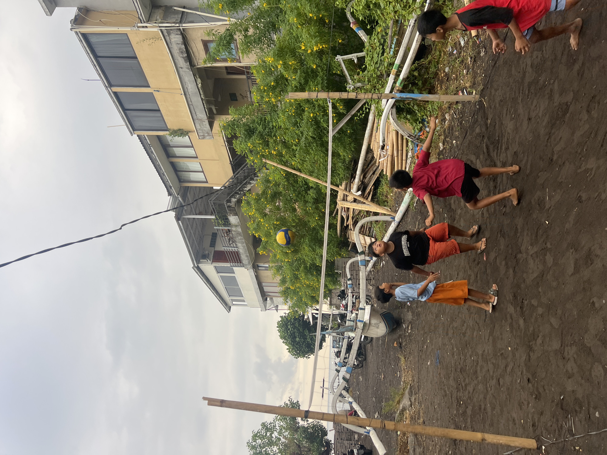 In Abang, Indonesia, on a cloudy day, four young boys are playing volleyball on a black sand beach, in front of a row of buildings and traditional outrigger boats. The volleyball net is strung between bamboo poles, and a yellow and blue volleyball is in the air. The buildings are multi-story structures with balconies and windows, and the surrounding vegetation is lush and green, with yellow flowers blooming. The beach is littered with wooden planks and other debris, suggesting it is a working area or a less maintained part of the coastline. The boys are actively engaged in the game, with one boy in the foreground reaching for the ball and others positioned on either side of the net. The scene conveys a sense of youthful energy and recreation amidst a coastal Indonesian environment.