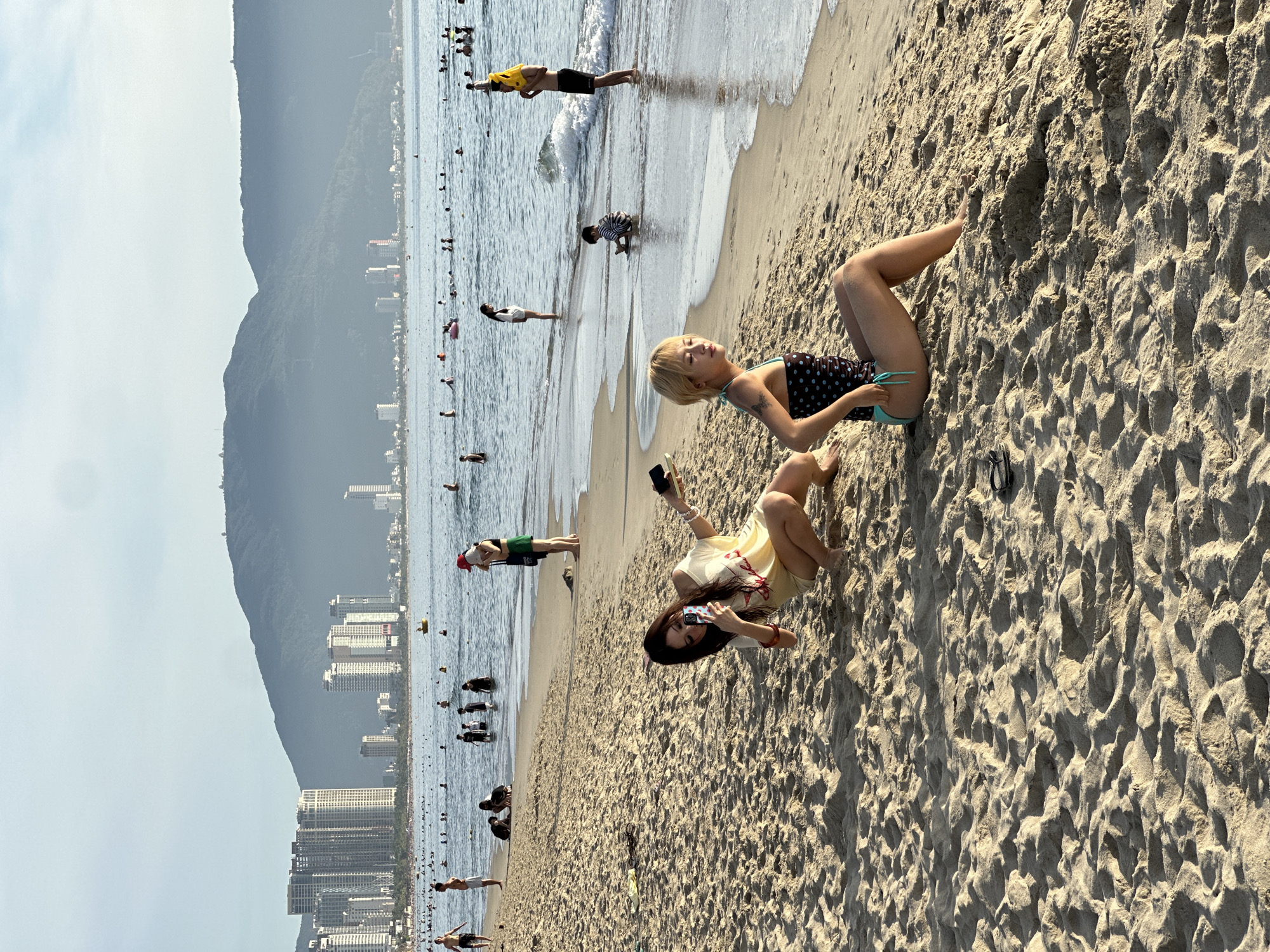 The image depicts a sunny day at a crowded beach in Đà Nẵng, Vietnam. The scene is filled with people enjoying the water and the sand. In the foreground, two young women are seated on the sandy beach. One, with blonde hair and a polka-dot swimsuit, is leaning back and appears to be holding a phone or camera. The other, with dark hair, is wearing a yellow top and shorts, and is actively taking a picture with her phone.
Further back, numerous people are scattered across the beach and in the shallow water. Some are standing, some are wading, and a few appear to be swimming or playing in the waves. A person in a yellow life vest is standing near the water's edge, facing away from the camera. The water is calm with gentle waves lapping the shore.
In the background, a range of tall buildings and a lush, green mountain form the backdrop of the beach. The clear sky suggests pleasant weather. The overall mood appears relaxed and festive, indicative of a typical beach day. The image captures the vibrant atmosphere of a popular tourist destination.