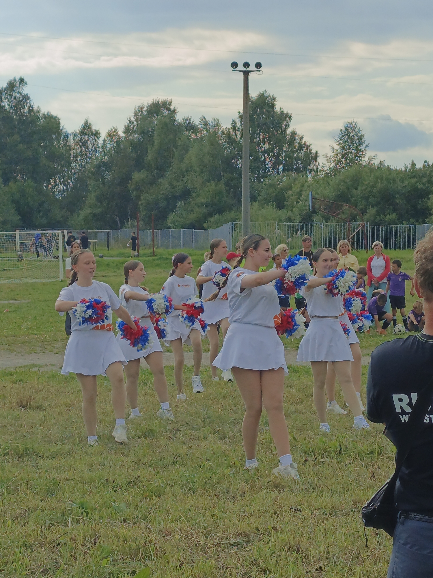 A group of young females in white cheerleader uniforms with red and blue pom-poms are performing a dance routine on a grassy field, likely a sports field, in Zlatoust, Russia. The background shows trees, a fence, and what appears to be a soccer goal. Several adults and children are observing the performance. The sky is overcast. A person in a black shirt is partially visible in the foreground.