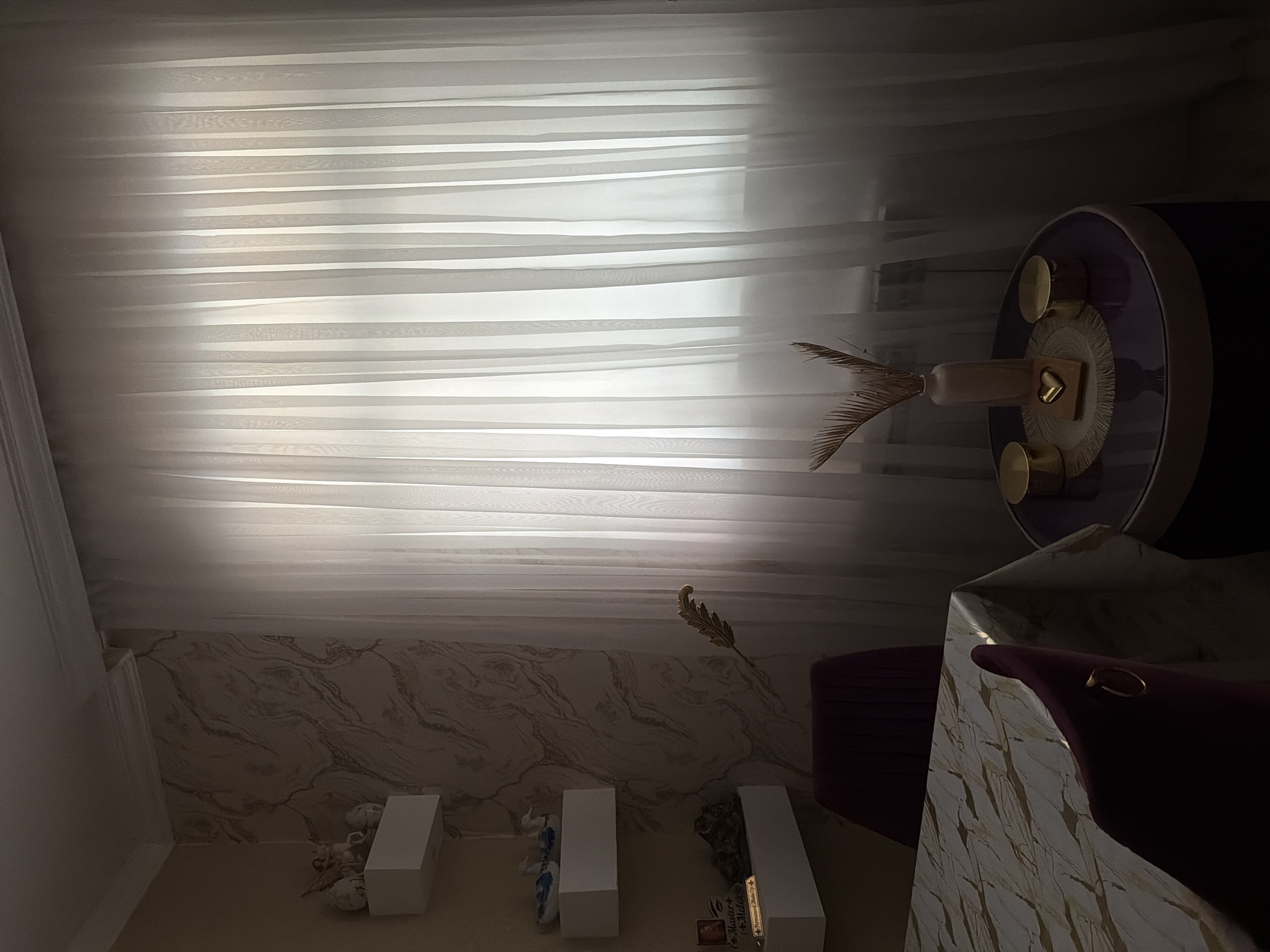 The image depicts an interior scene, likely a dining area or living room, with a focus on a window covered by sheer white curtains. To the left of the window, on a wall with marbled wallpaper, are three white shelves. The top shelf holds two small figurines, possibly angels. The middle shelf contains two blue and white ceramic objects. The bottom shelf displays a framed picture with text, a small stone or decorative object, and a gold-colored feather-like ornament. In the foreground, a table is partially visible, covered with a tablecloth featuring a marble-like pattern in white and gold. Two dark red chairs with gold rings on the back are positioned around the table. To the right of the table, a small round table with a purple top holds a vase with dried foliage, a heart-shaped ornament on a coaster, and two gold cylindrical objects. The lighting is dim, suggesting it is either early morning or late evening. The image does not contain any explicit indicators of the city of Digah, Azerbaijan, or any identifiable individuals.