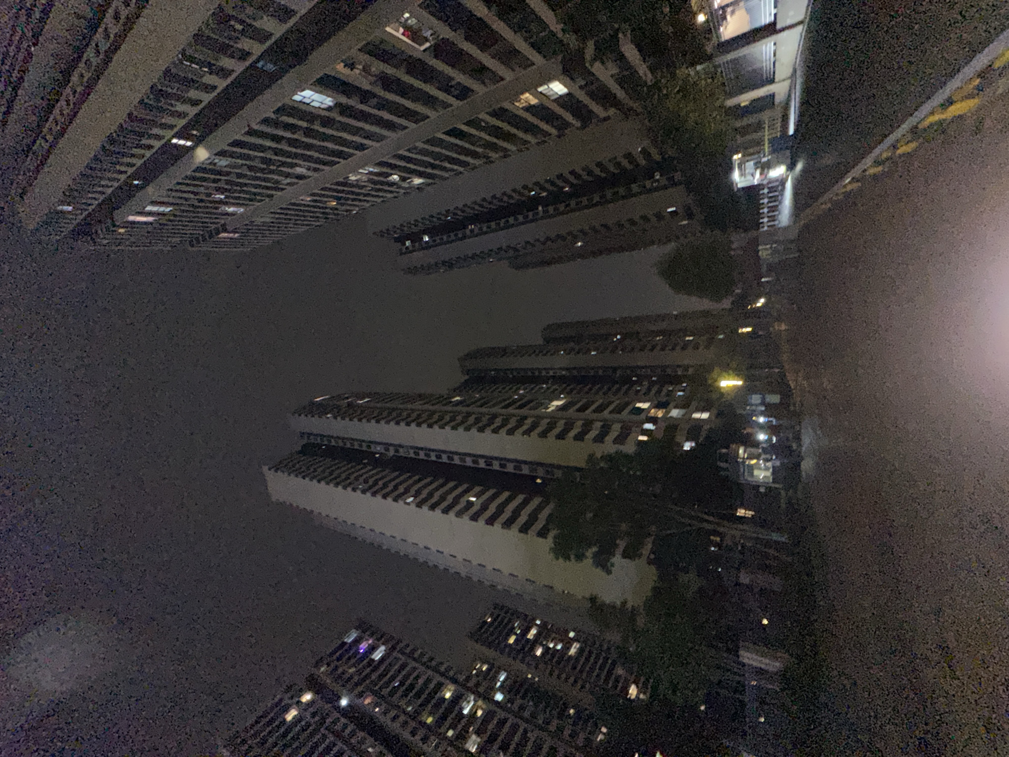 The image depicts a nighttime scene in what appears to be a densely populated urban area. The view is oriented upwards, showcasing a cluster of tall buildings with numerous illuminated windows. The buildings are uniformly structured, suggesting residential or commercial complexes. The environment is dimly lit, with only the building lights illuminating the scene. Some foliage is visible interspersed among the buildings. The location is specified as the Chongqing High-tech Zone in China. There are no people visible in the image.