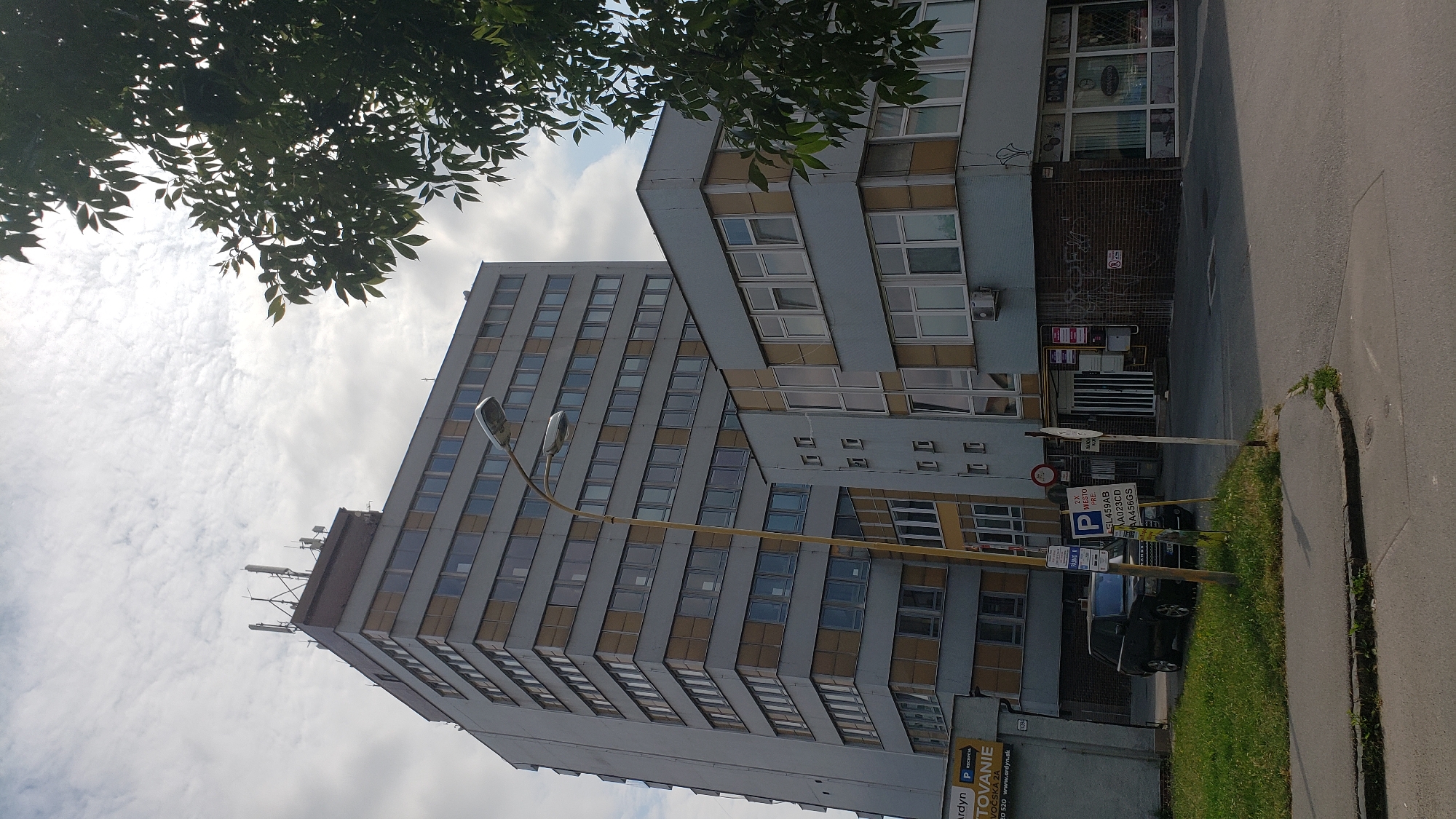 This is a daytime, medium-angle exterior shot of a multi-story office building and a smaller attached structure, located in the city of Prešov, Slovakia. The buildings are modern, grey, and appear to be in a commercial or industrial district. The larger building is tall and rectangular with numerous windows and several antennae on its roof. The attached structure is shorter and also features many windows, with a slightly different architectural style. A street lamp with two lights stands in front of the buildings. In the foreground, a black SUV is parked on the side of a road next to a grassy area. There are several signs visible, including parking signs and a sign for "ARSYN." A tree with green leaves is partially obscuring the right side of the frame. The sky is cloudy. There are no people visible in the image.