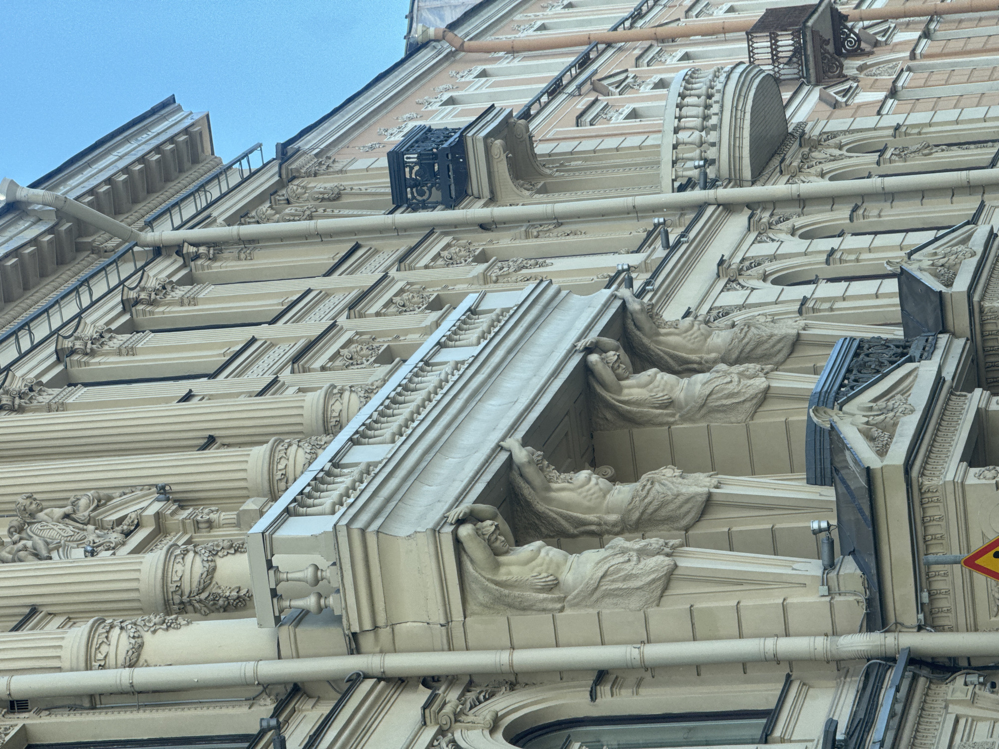 This is a low-angle, close-up shot of the ornate facade of a building in Saint Petersburg, Russia. The architecture is characterized by intricate detailing and classical motifs, predominantly in a light beige stone. The building features multiple levels with repeating patterns of pilasters, cornices, and decorative friezes. Prominently displayed on several levels are sculpted figures of reclining nude women, appearing to support architectural elements. These figures are rendered in a classical style, with draped fabric and detailed musculature.
Above these figures, there are horizontal pipes running across the facade, likely part of the building's drainage system. Further up, there are balconies with decorative ironwork, and small, dark window-like structures jutting out from the building. The sky above is a clear, pale blue, suggesting daytime with fair weather. The overall impression is one of historical grandeur and artistic craftsmanship, typical of the architecture found in Saint Petersburg. No visible text is present in the image.