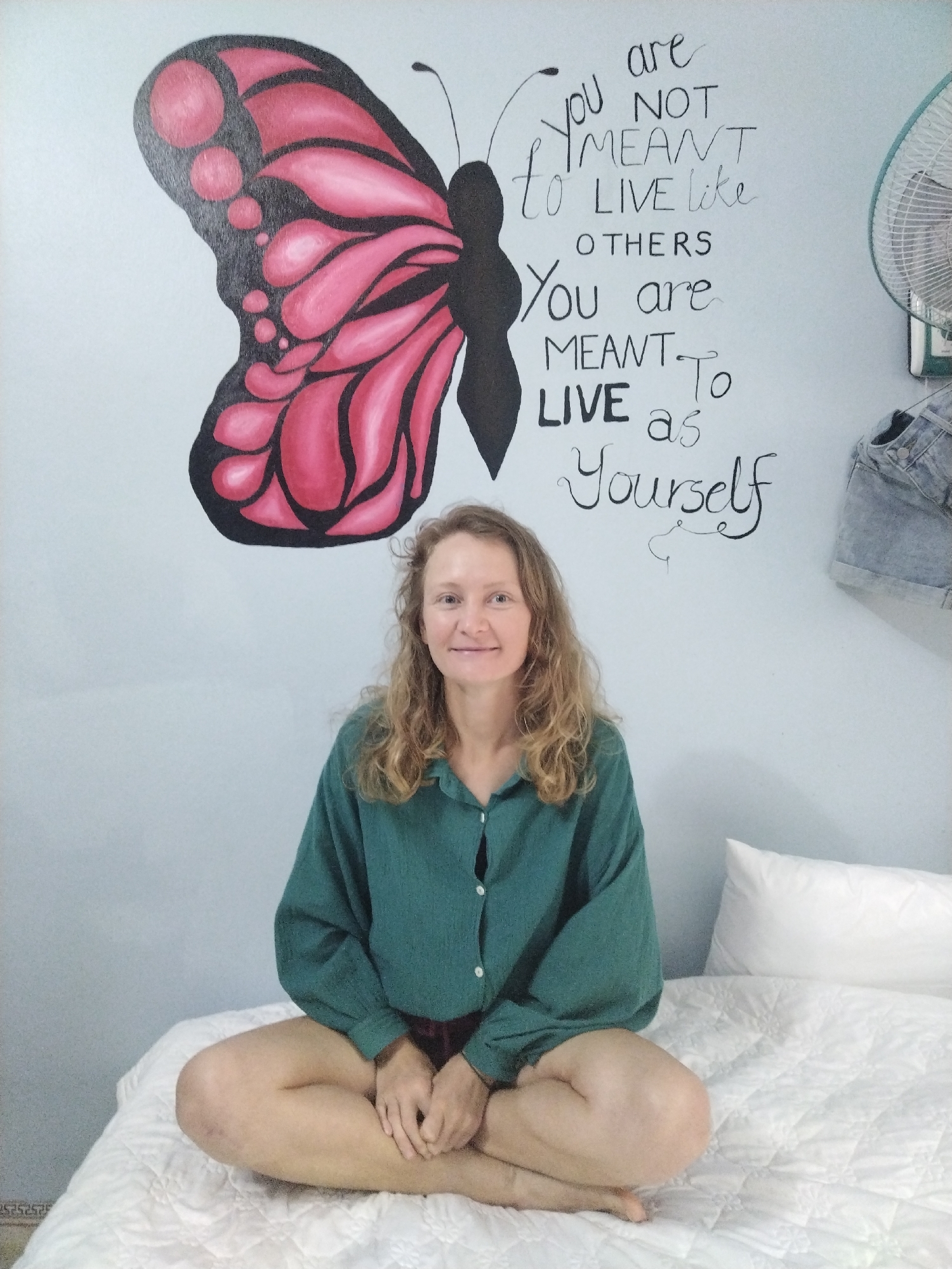 A woman with light, wavy blonde hair, wearing a loose-fitting dark green long-sleeved shirt and dark shorts, sits cross-legged on a white quilted bed. She is looking directly at the camera with a calm, friendly expression and a slight smile.
The setting is an indoor space, likely a bedroom, with a light blue or pale grey wall as the background. Dominating the wall to the left is a large, vibrant mural of a butterfly with pink wings outlined in black, and a black body. To the right of the butterfly, written in black script and block letters, is the inspirational text: "You are NOT MEANT to LIVE like OTHERS You are MEANT to LIVE as Yourself".
Further to the right on the wall, an oscillating white fan is mounted high, and below it, a pair of light-washed denim shorts hangs. The lighting is bright and even, suggesting daytime. A white pillow is visible on the bed to the woman's right. The overall atmosphere is relaxed and personal. The setting is identified as Bai Chay Ward, Vietnam.