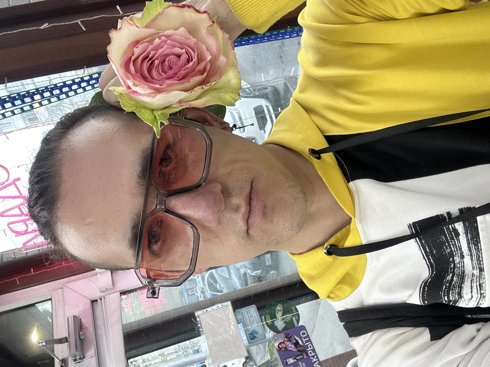 A vertically oriented, close-up image captures an adult male posing for a selfie, looking directly at the camera with a neutral expression. He has dark, pulled-back hair with a visible receding hairline. He wears large, hexagonal eyeglasses featuring translucent pink-orange tinted lenses and clear frames. A vibrant, large rose, primarily pink with yellow and green accents, is prominently tucked behind his right ear, serving as an adornment.
The man is dressed in a bright yellow hoodie, which transitions into a distinct black and white patterned section on the lower portion, adorned with a black drawstring. The background suggests an indoor setting, likely near a window, offering a blurred glimpse of an urban environment outside. Faint outlines of buildings and a white vehicle are discernible through the glass. Various items, including cards and papers, are displayed on or behind the window.
Visible Russian text on these items includes "ЗАКРЫТО" (CLOSED) on a purple card, and partially visible words like "САРАТОВ" (SARATOV) and "НА ВЫБОР" (FOR CHOICE) on other cards. The natural light indicates it is daytime. The setting, potentially a shop or cafe, is located in Saratov, Russia.