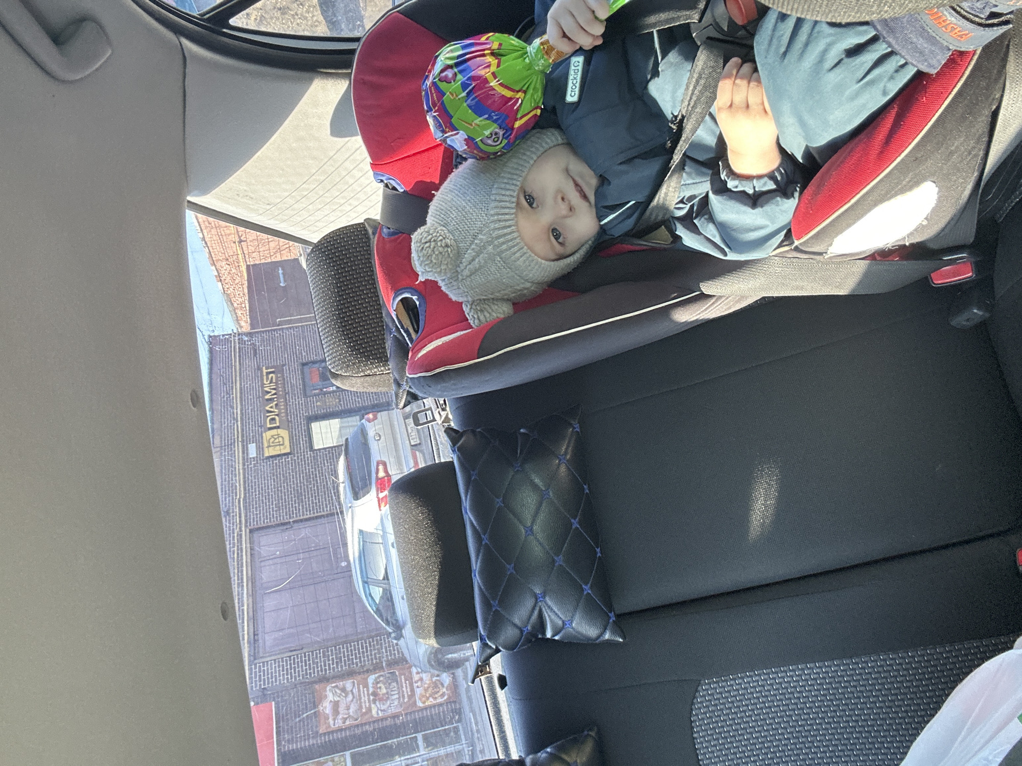 The image depicts the interior of a vehicle, specifically the rear passenger compartment, with an external view through the windows. The scene is located in Chaikovsky, Russia, outside a building identified by a visible sign as "DIA.MIST SOURCE HOOKAH," likely at ulitsa Lenina, 67A.
Inside the vehicle, a young child, appearing to be a toddler, occupies a red and black child safety seat positioned on the right side of the rear bench. The child wears a grey knit hat with two pom-poms and a dark teal/blue winter jacket bearing the "crockid" brand logo. They are secured by a black harness and hold a large, multi-colored lollipop, wrapped in clear plastic, in their right hand. The child is facing slightly left, towards the camera, with a neutral expression. The vehicle's rear seats are dark fabric, with a black quilted headrest pillow with blue stitching visible on the left rear seat. The interior roof lining is light grey.
Through the rear passenger window, a dark brick commercial building is visible. This building features a large, dark metal double-door, likely a garage or loading bay, and several windows. A prominent yellow sign with black lettering displays "DIA.MIST" at the top and "SOURCE HOOKAH" below. Advertisements, possibly for food items, are partially visible on some of the building's windows. A silver SUV, possibly a Kia model, is parked in front of the building, with its rear facing the vehicle from which the photograph was taken. The SUV's license plate shows partial registration "877MA." The sky appears clear, and strong sunlight suggests daytime conditions.