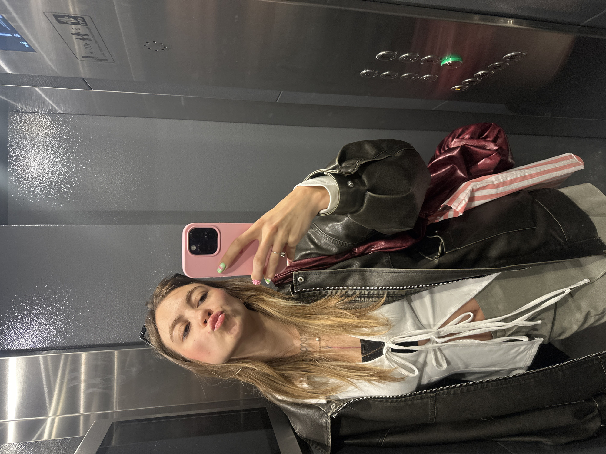 A young woman with long, blonde hair is taking a selfie in an elevator. She is wearing a black leather jacket over a white top and grey pants. She is holding a pink phone in her right hand, and her left hand is resting on the phone. She is pouting her lips and looking at the camera. The elevator is made of stainless steel, and the walls are grey. There is a panel of buttons on the right side of the elevator, and the number 7 is lit up. The woman is wearing rings on her fingers and a necklace around her neck. Her nails are painted green with a floral design.