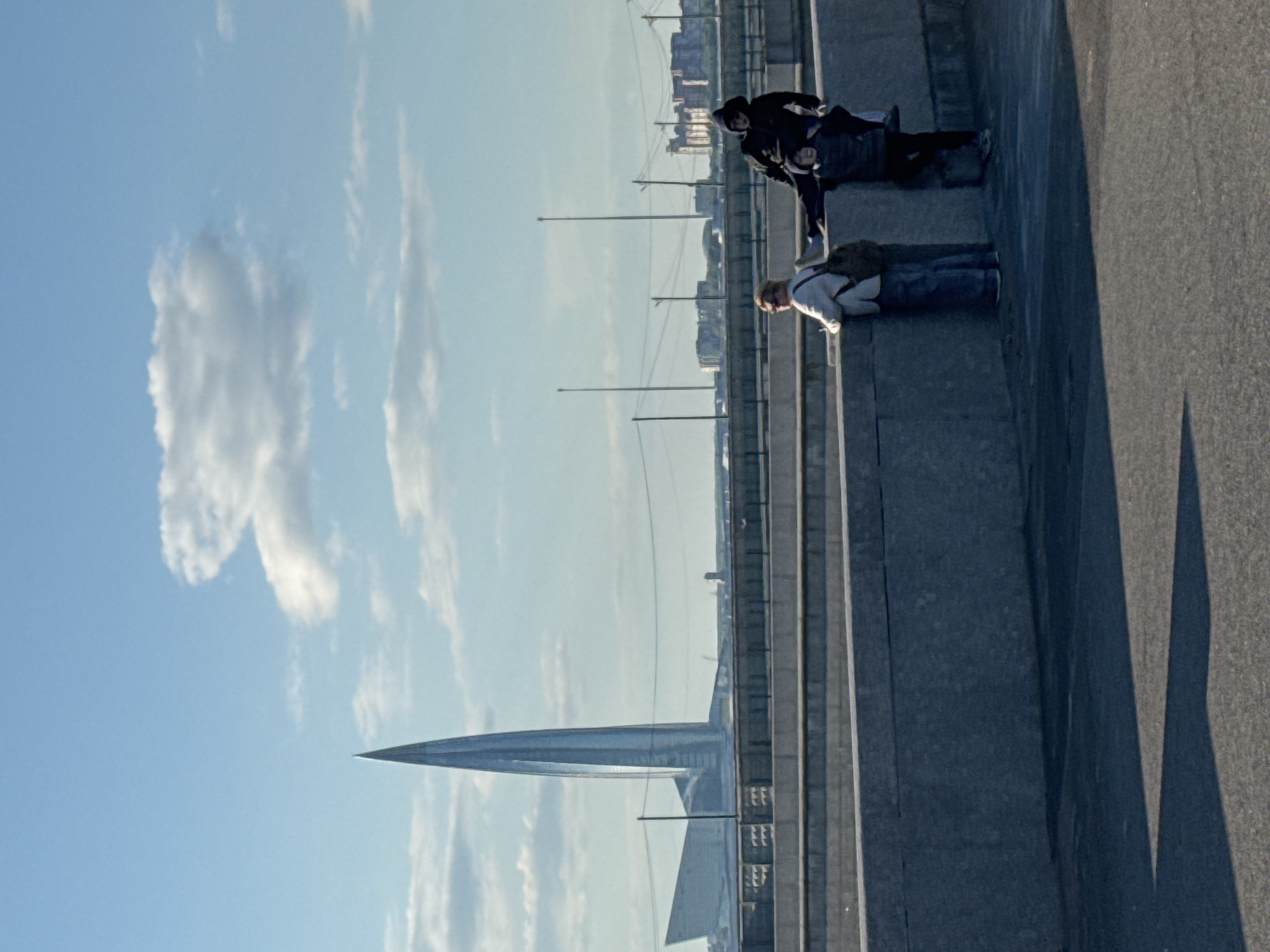 The image shows a view of the Lakhta Center skyscraper in Saint Petersburg, Russia, from an elevated vantage point, possibly a bridge. Two people are lying or sitting on a concrete structure in the foreground. The sky is blue with scattered clouds.