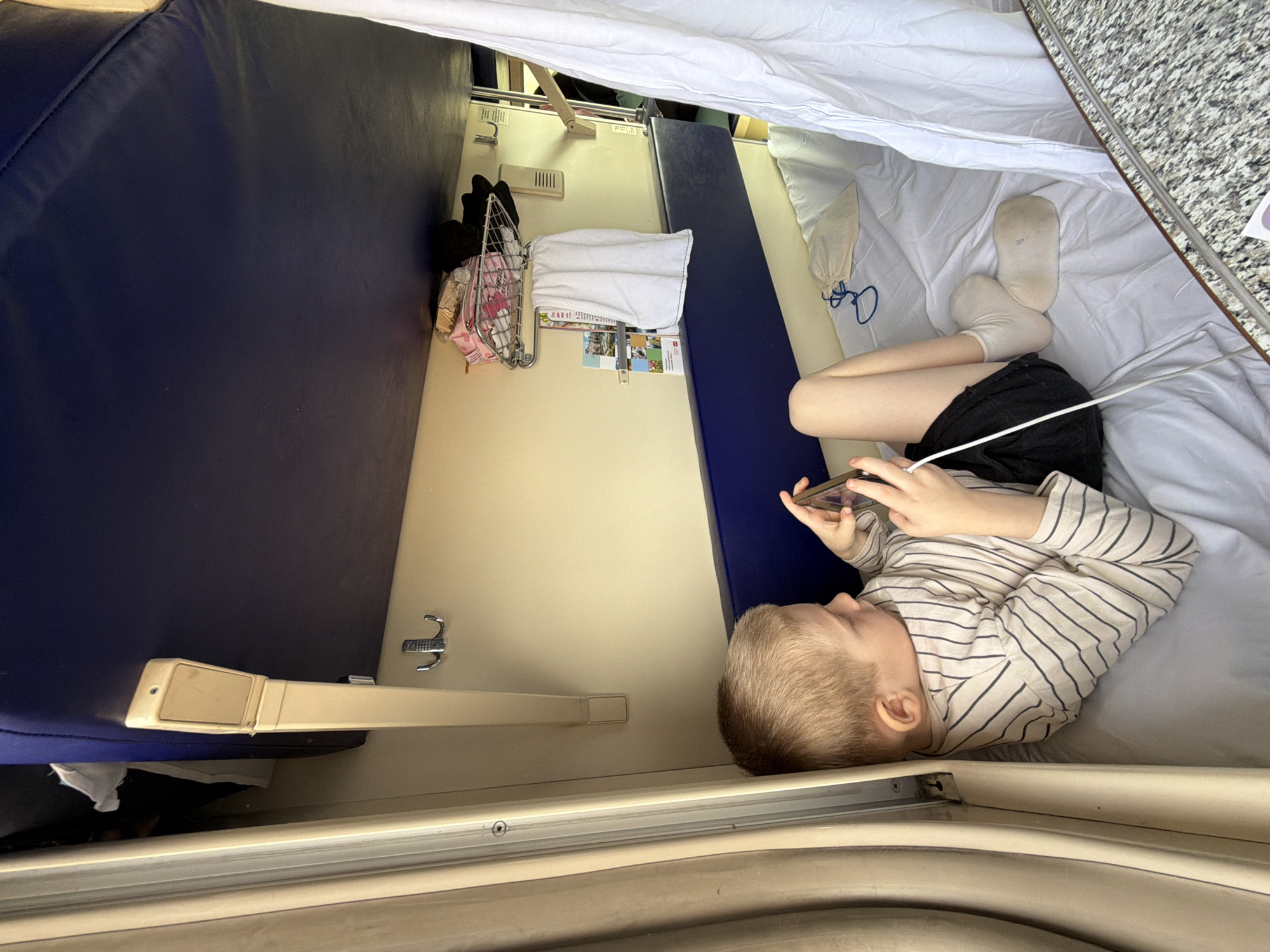 A young child, likely a boy, lies on a bed inside what appears to be a train compartment. He is wearing a striped shirt and dark shorts, and is holding a smartphone in his hands. The bed has white sheets. A second berth with dark blue padding is visible on the left. A white sheet is draped over the top of the compartment, possibly to create a sense of privacy. A small metal rack with personal items hangs on the wall. The location, Medvyodovskaya, Russia, is assumed due to the query requirements.