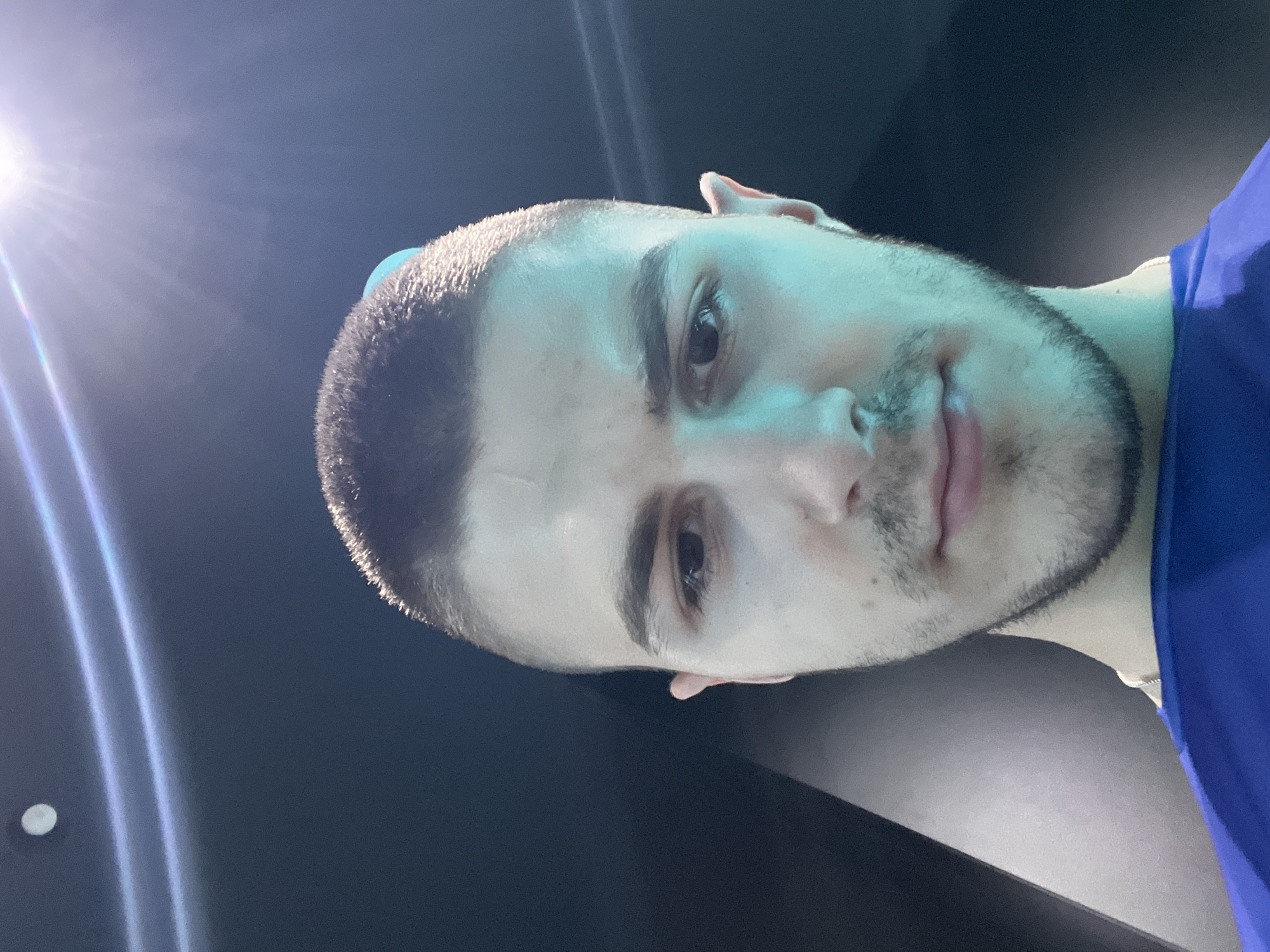 The image is a close-up, rotated portrait of a young man with short, dark hair and a light beard. He is wearing a blue shirt. The background appears to be dark, possibly a wall or ceiling, with some light fixtures visible on the left side. The overall setting suggests an indoor location, but the specific location in the city of Unknown, Unknown, cannot be determined from the image.