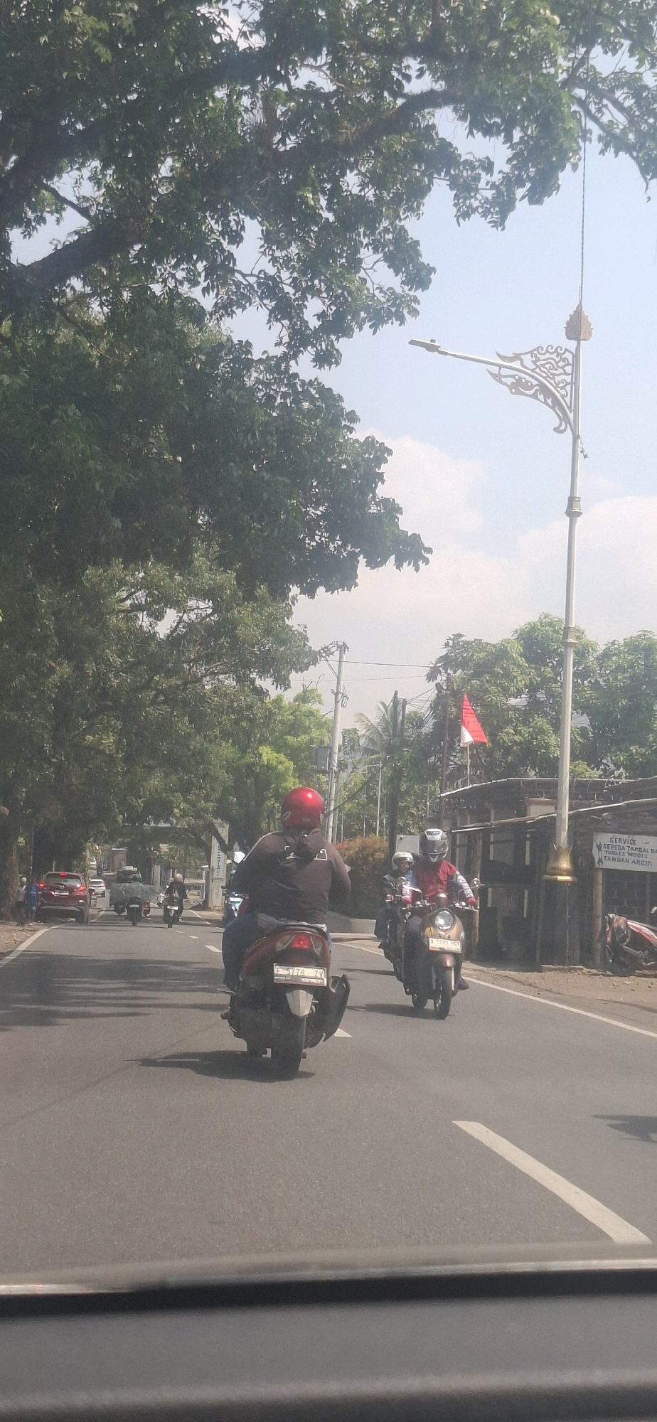 The image depicts a street scene in Sukasari, Indonesia, under a bright, clear sky. The foreground shows the interior of a vehicle, likely a car, with a dark dashboard visible at the bottom. The main focus is the road ahead, with several motorcycles and a car driving on it.
In the center of the road, a person wearing a red helmet and a dark jacket is riding a scooter. The scooter has a white license plate with the inscription "F 778 ZV". To the right of this scooter, another scooter is visible, carrying two people. The rider wears a red jacket and a white helmet, and the passenger has a white helmet. This scooter has a yellow license plate that reads "D 1335 PCY".
Further down the road, more motorcycles are seen. On the left side of the road, a red car is parked. The road is lined with trees on both sides. On the right side of the road, there is a building with a sign that is partially visible and appears to be in Indonesian. A red and white flag, likely the Indonesian flag, is flying on a pole. A decorative street lamp with intricate metalwork stands tall near the flag. The overall environment suggests a typical daytime street in a city or town.