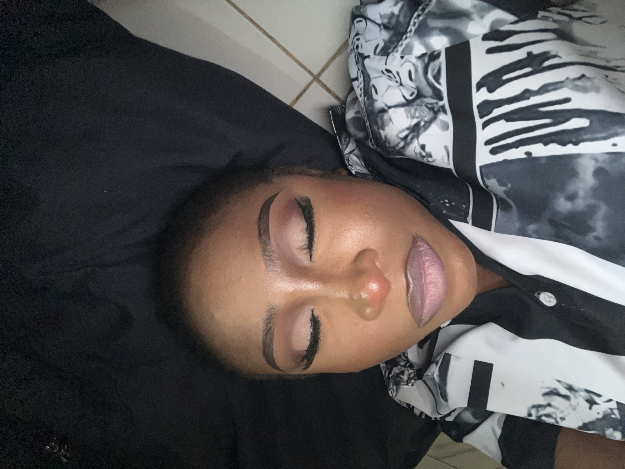A medium shot captures a close-up of a woman lying down, with her eyes closed. She is wearing a black and white patterned shirt. Her lips are painted a light purple color, and her eyebrows are sharply defined. The background is a dark, solid color, possibly fabric, and a section of tiled flooring is visible at the bottom left. The scene appears to be indoors, and given the context of Abuja, Nigeria, it could be a personal residence or a beauty salon.