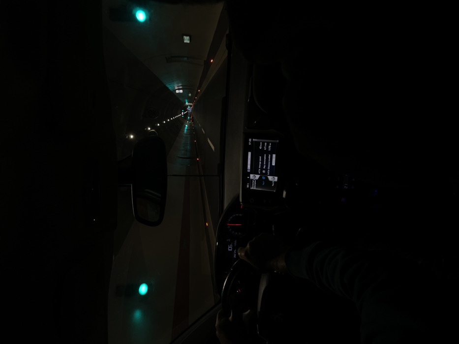 The scene depicts the interior of a vehicle moving through a tunnel in Mateševo, Montenegro. The perspective is from the front passenger compartment, looking forward through the windshield.
A driver's hands are positioned on a dark steering wheel. The instrument cluster shows "106" prominently, indicating speed, and "266.2km" for a trip distance. A central infotainment screen displays "18°C," the time "17:02," an average speed of "42 Km/h," an average fuel consumption of "4.7l/100," and a total calculated distance traveled of "1175 km." A dark rearview mirror is suspended from the ceiling. The indistinct, dark form of a second occupant is visible in the right foreground, consistent with a front passenger.
The tunnel interior features light-colored walls with a continuous red horizontal stripe at a lower level. The tunnel ceiling contains an array of recessed white lights extending into the distance. Large, circular green lights are affixed to both the left and right tunnel walls at regular intervals. The asphalt roadway comprises two lanes delineated by white markings. Small, orange-red reflective markers are present along the road's edges. The tunnel structure recedes into the background, maintaining consistent artificial illumination.