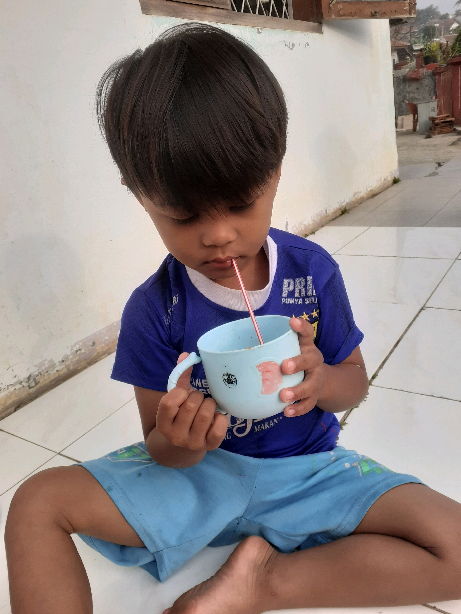 A young child, likely male, with dark hair is sitting on a tiled outdoor floor, possibly a patio or walkway, and drinking from a light blue cup with a pink straw. The child is wearing a blue t-shirt with white trim and blue shorts with some light green graphics. The child's head is bowed, and their eyes are focused on the cup. The background shows a white wall, a wooden window frame, and glimpses of houses and vegetation, suggesting a residential area. The overall scene appears to be in a warm climate. Based on the provided information, this image was taken in the city of Sumedang, Indonesia.