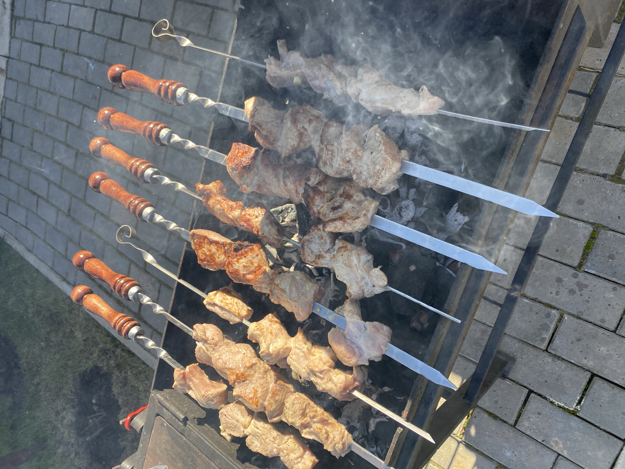 The image shows six metal skewers with wooden handles holding pieces of meat cooking over a charcoal grill outdoors. The meat on some skewers is visibly more cooked than on others. Smoke rises from the grill. The grill sits on a paved surface made of rectangular gray bricks. The location is Гаревая, Russia.