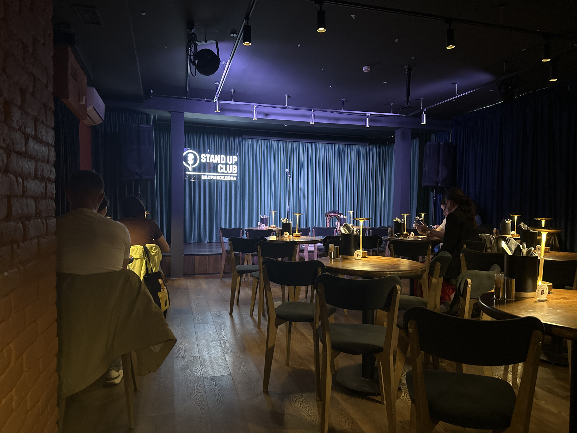 The image captures the interior of a dimly lit comedy club named "Stand Up Club" located at "Nagriboedova" (Griboyedov Canal) in Saint Petersburg, Russia. The room is set up for a performance with a stage at the far end draped with blue curtains and a neon sign that reads "Stand Up Club На Грибоедова". Multiple round wooden tables are arranged in front of the stage, each illuminated by a small table lamp and some with containers that appear to hold napkins and possibly utensils. Several people are seated at the tables, some facing away from the camera. The room is lit by spotlights on tracks and general ambient lighting. The walls are dark, with a brick section on the left. The floor is made of wooden planks.