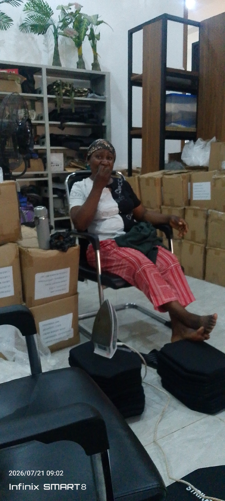 In Kakau, Nigeria, a woman is seated in a black office chair, holding her hand to her mouth in surprise. She wears a white and black patterned shirt and red and white striped pants. Her feet are bare. In front of her, on the floor, is a black ironing board with an iron on it. The room appears to be a storage area, with shelves filled with items and stacked cardboard boxes behind the woman. Artificial plants are visible on a shelf in the background. The overall lighting suggests an indoor environment, possibly a warehouse or a commercial space. The timestamp at the bottom indicates the image was taken on July 21, 2026, at 09:02.