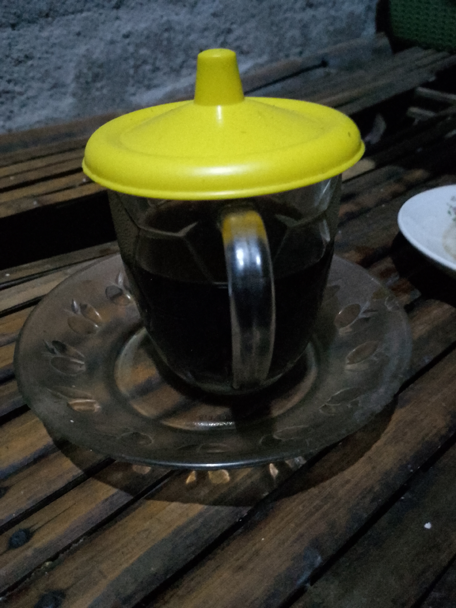 A close-up shot captures a glass of dark liquid, likely coffee, topped with a bright yellow lid. The glass sits on a transparent, patterned saucer, which rests on a wooden surface constructed from horizontal planks. To the right of the glass, a white plate with a faint floral pattern is partially visible. The background consists of a textured, grey wall and a dark, woven material. The lighting suggests an indoor setting, possibly in Kauman, Indonesia, given the context.
