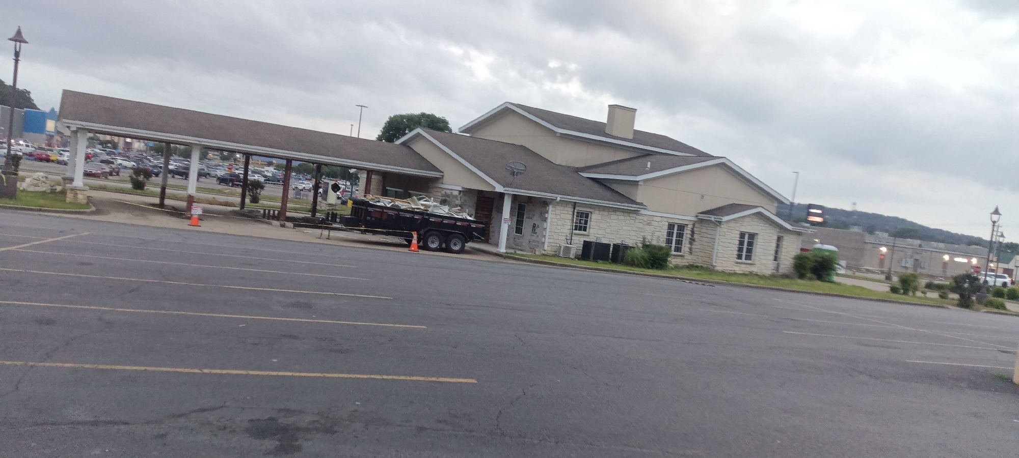 The image shows a parking lot in front of a single-story commercial building. The building has a combination of tan-colored siding and stone facade, a gabled roof with a chimney, and several windows. A covered walkway supported by pillars extends from the building towards a large parking area filled with vehicles. A black trailer filled with debris is parked near the walkway, marked by orange traffic cones. The parking lot is asphalt with yellow lane markings. In the background, there is a distant hill and additional buildings. The sky is overcast, suggesting a cloudy day. The location is Van Buren, USA.